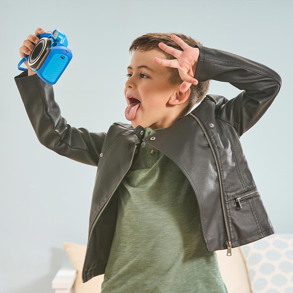 Ein junger Junge, der eine blaue Kamera hält, Grimassen schneidet, eine Jacke und ein grünes Hemd trägt.