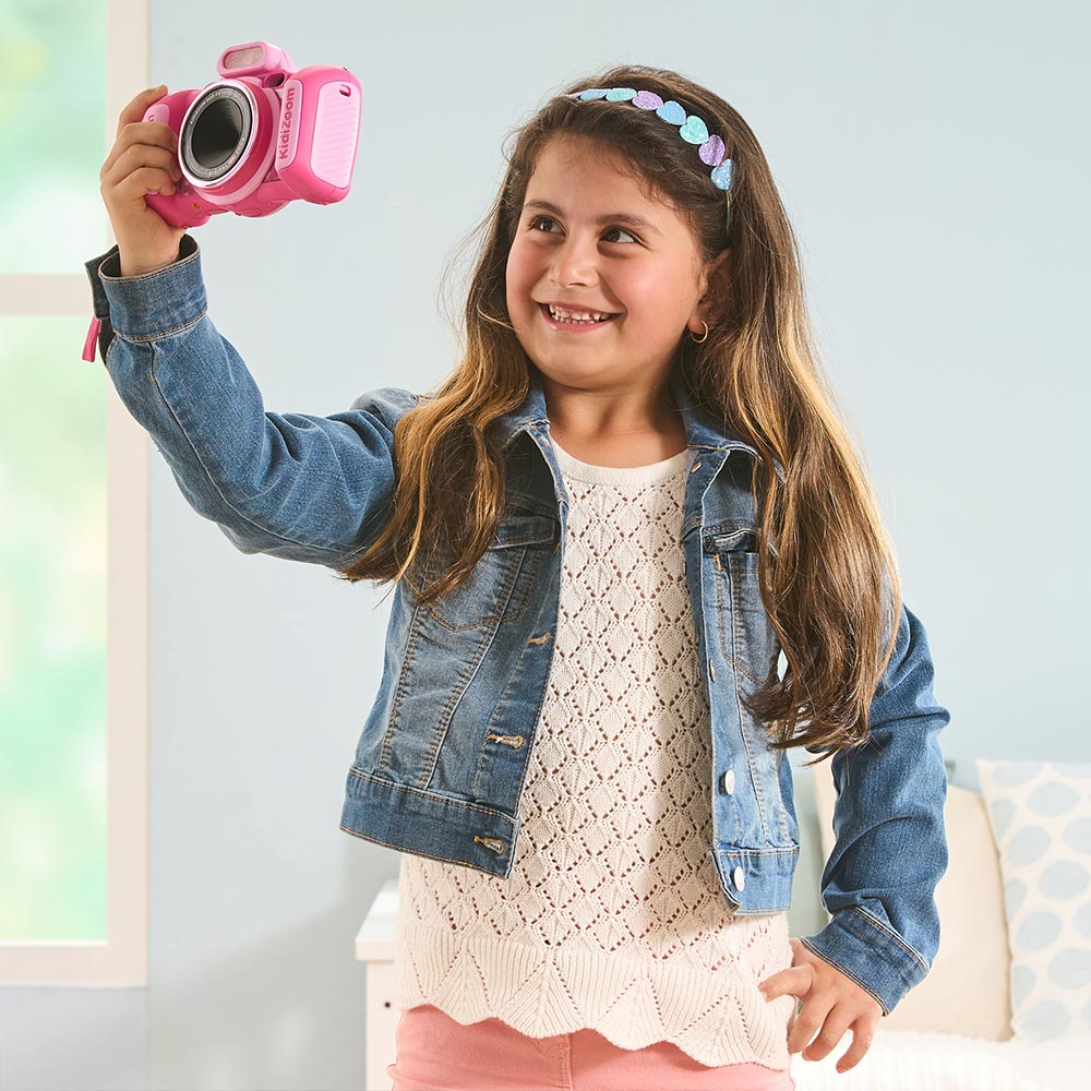 Una ragazza che tiene una macchina fotografica rosa, indossa una giacca di jeans e sorride.