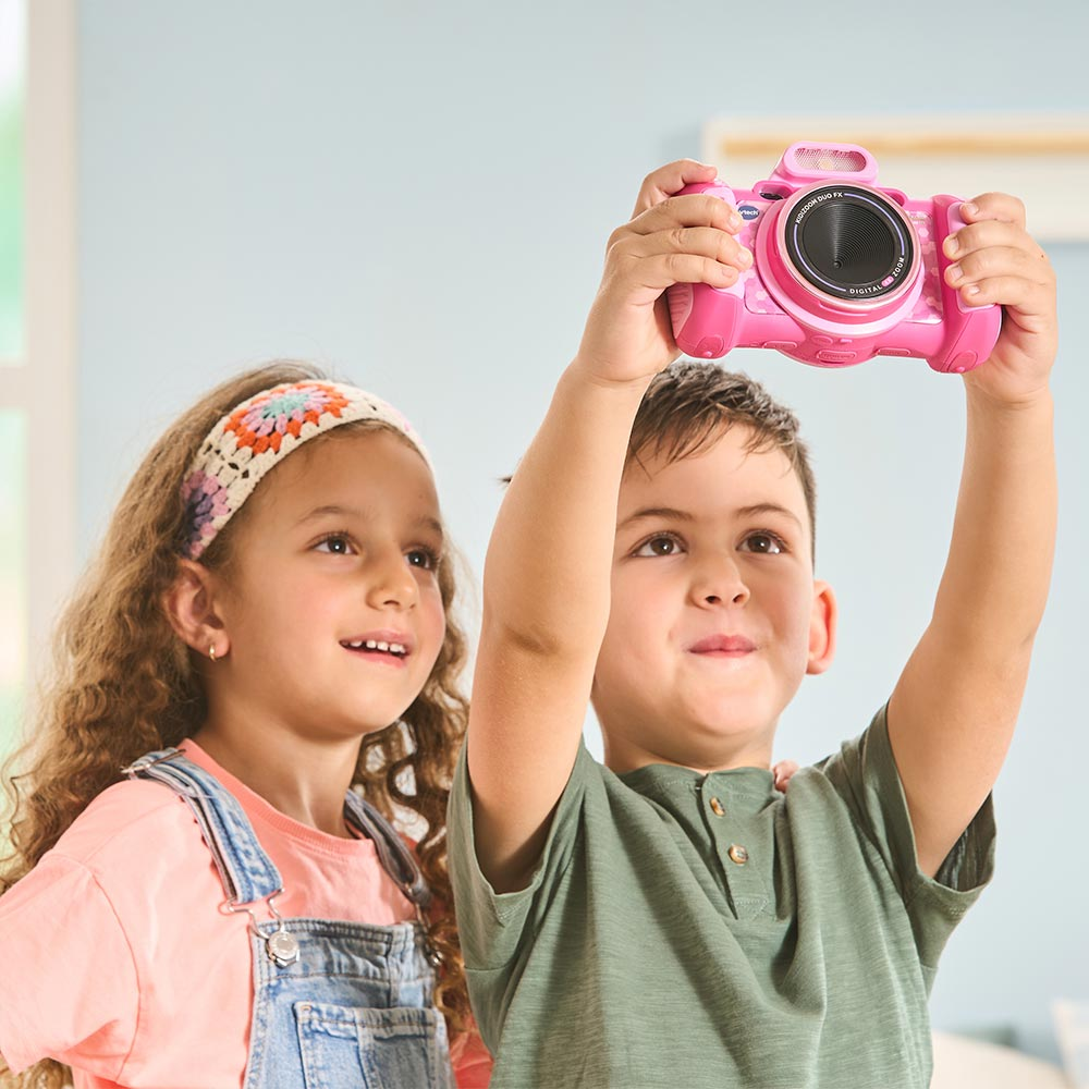 Due bambini che tengono in mano una fotocamera rosa, sorridendo. Uno la tiene in alto, l'altro la guarda. Interno.