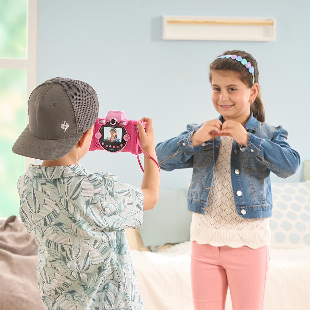 Un ragazzo fa foto con una fotocamera rosa. Una ragazza posa. Lo sfondo è una stanza con finestra e letto.