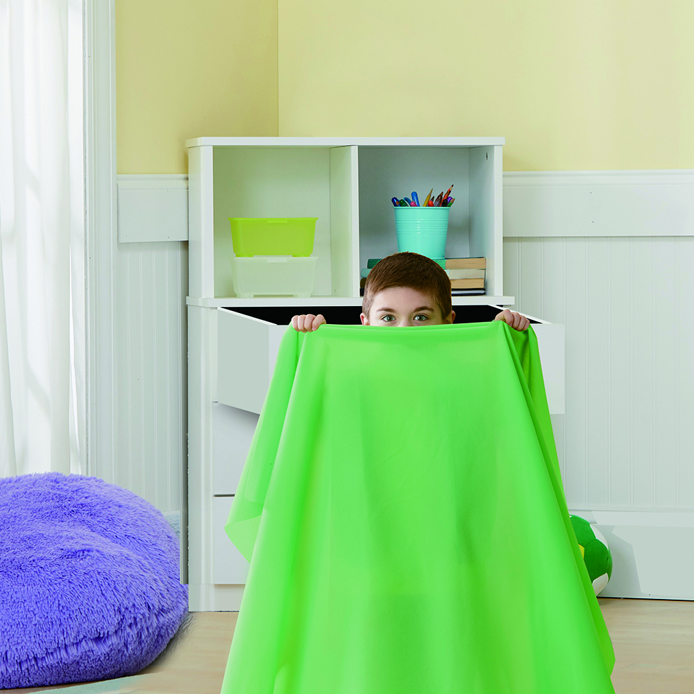 Un enfant regarde par-dessus un tissu vert dans une pièce avec un pouf violet et une étagère.