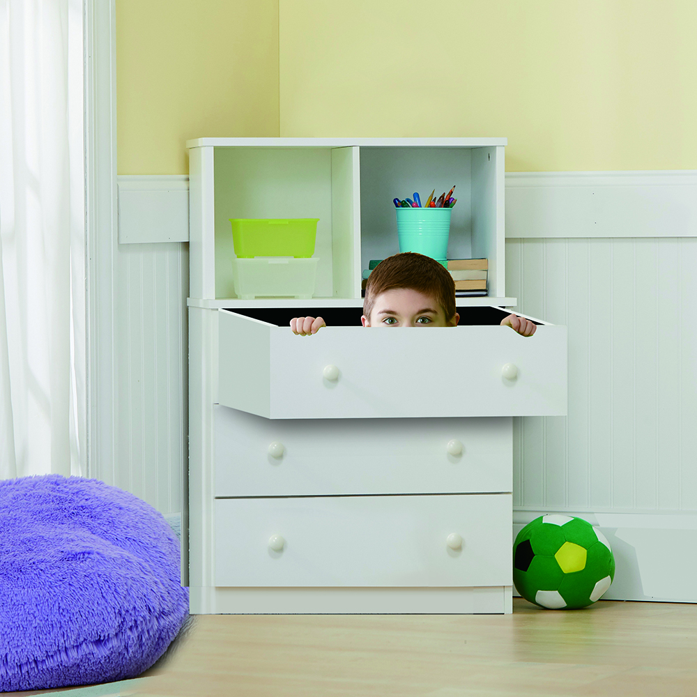 Un garçon regardant depuis un tiroir. Une commode avec des tiroirs est dans une pièce. Un pouf violet est au sol.