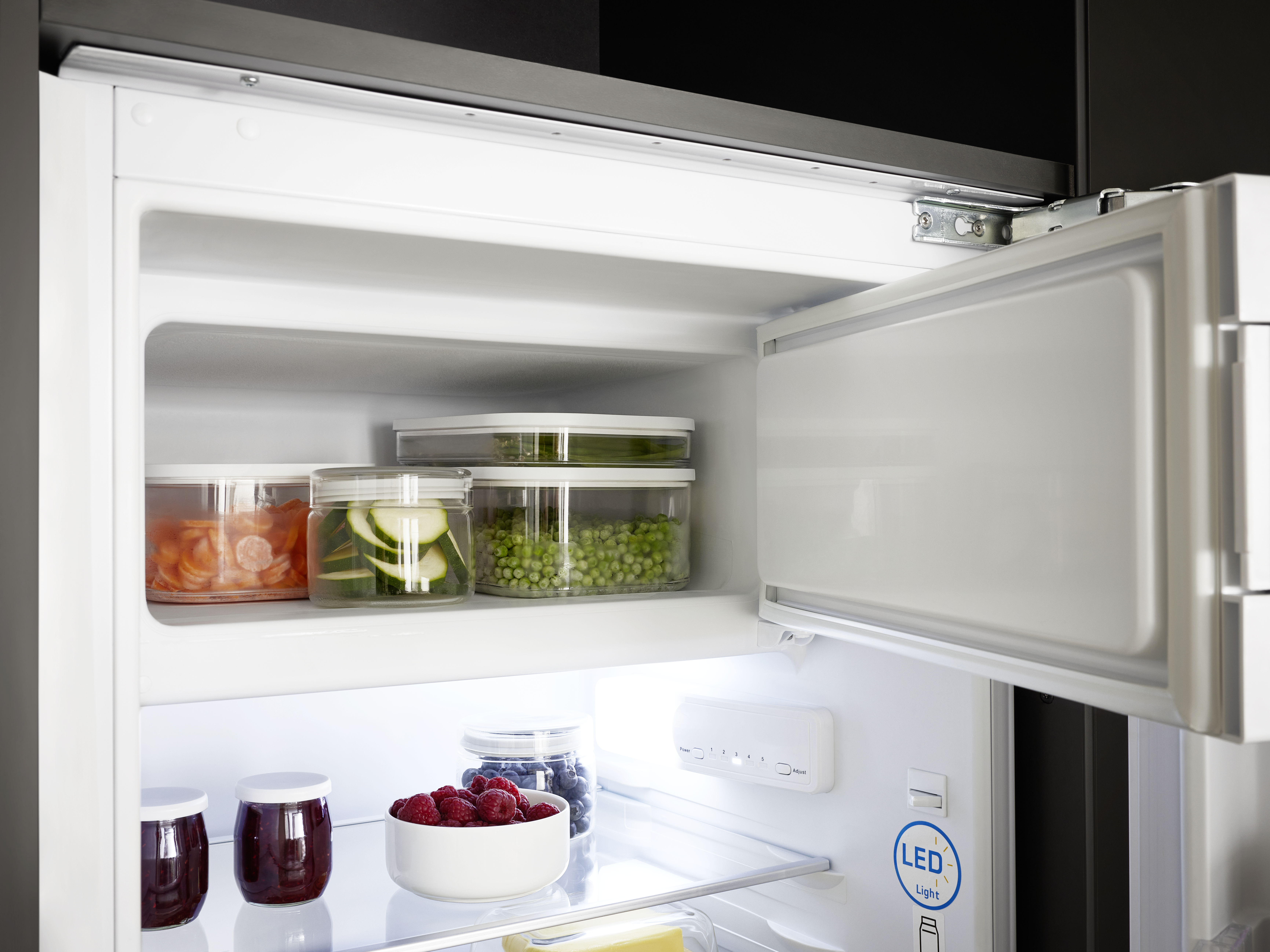 Offener Kühlschrank mit Regalen voller Lebensmittel. Gläser mit Marmelade, Gemüsebehälter und Himbeeren.