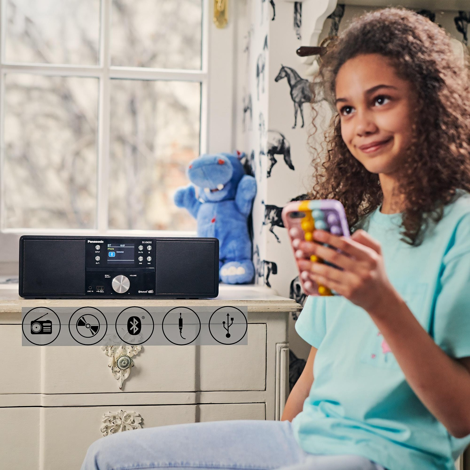Ragazza con telefono, ascolta un altoparlante Panasonic. Vengono mostrate icone per radio, CD, Bluetooth, jack e USB.