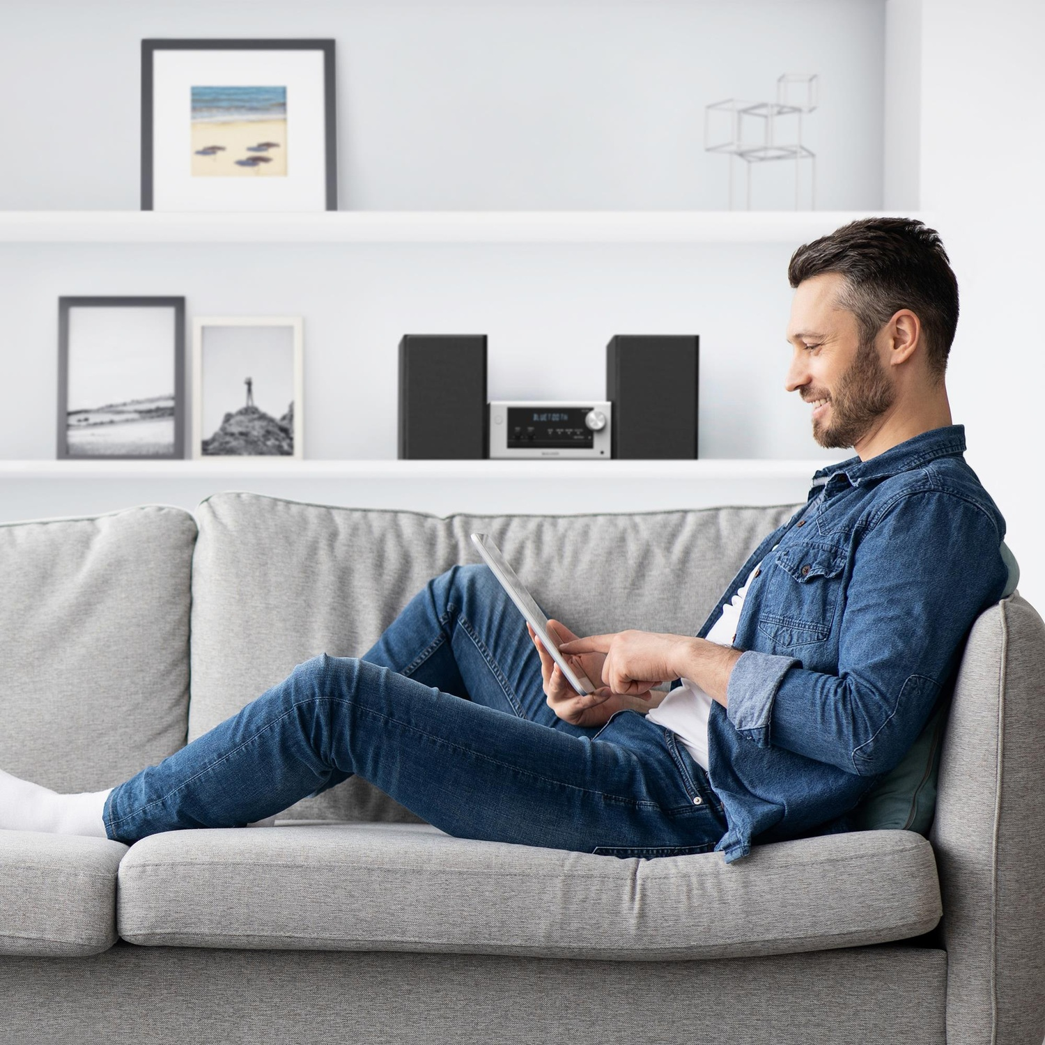 Uomo sul divano grigio usa tablet. Sfondo: mensole, foto, impianto stereo.