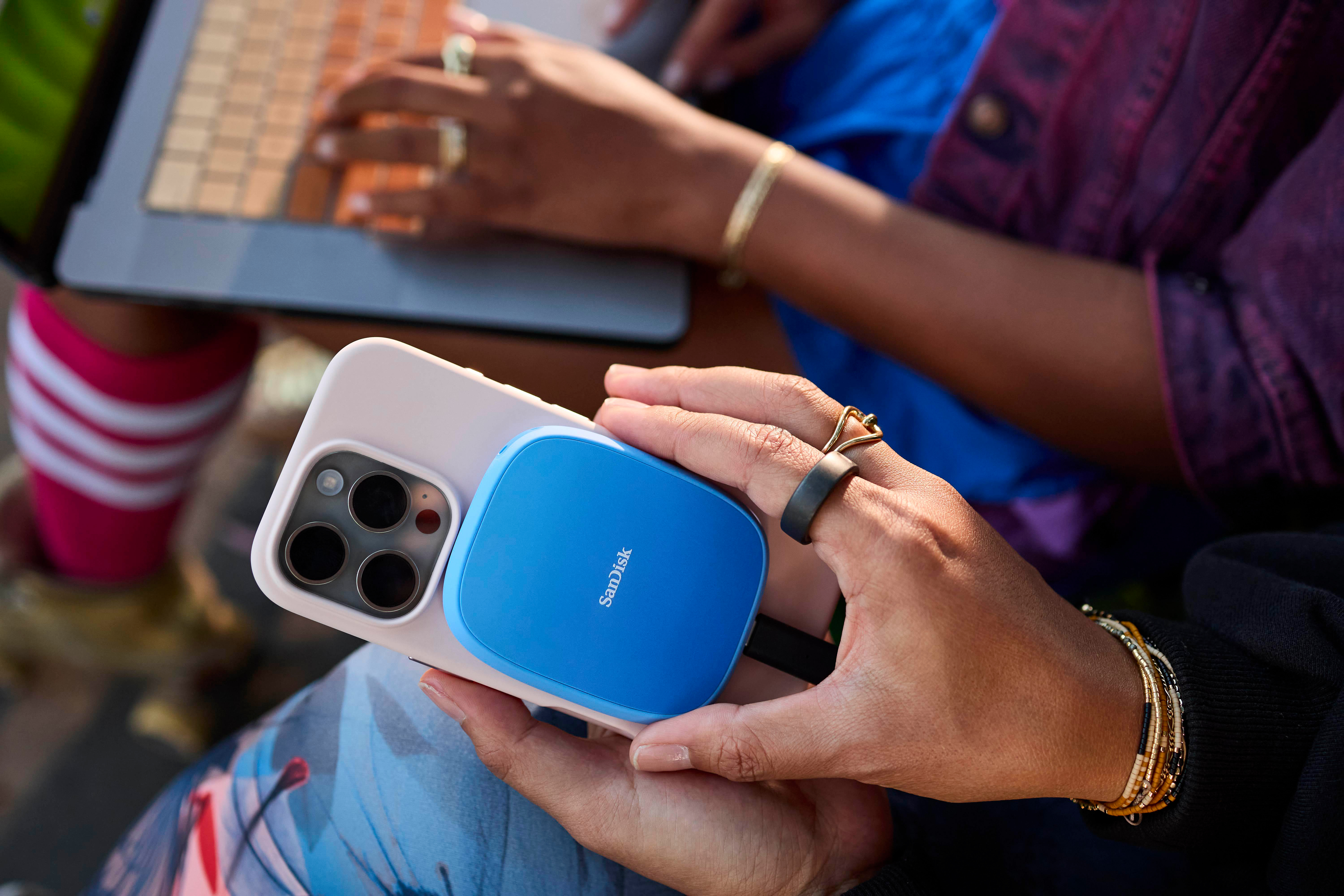 Eine Person hält ein Telefon mit einem blauen Sandisk-Gerät. Eine andere Person tippt auf einem Laptop. Der Hintergrund ist unscharf.