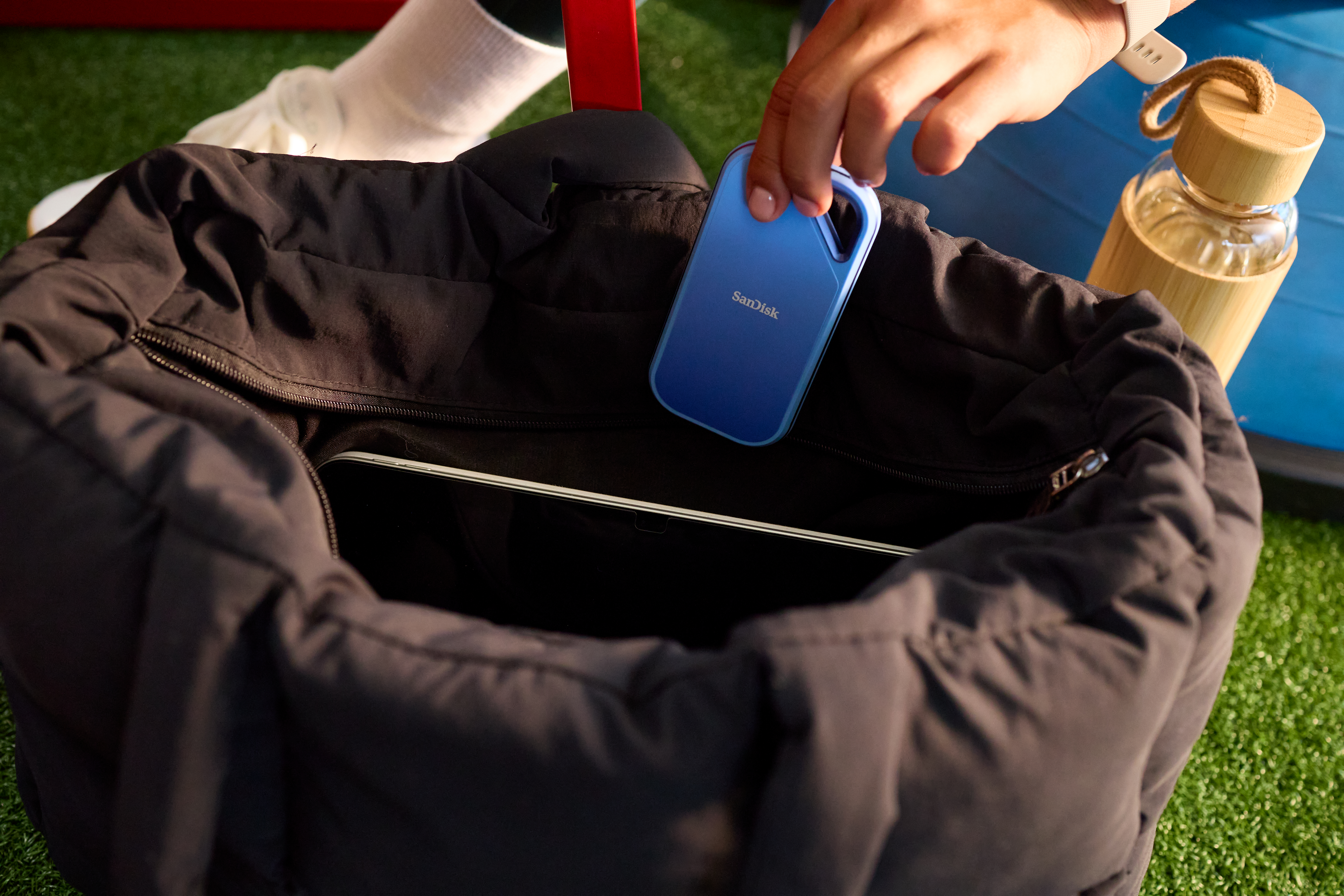 Eine Hand legt eine blaue SanDisk externe Festplatte in eine schwarze Tragetasche. Grünes Gras im Hintergrund.