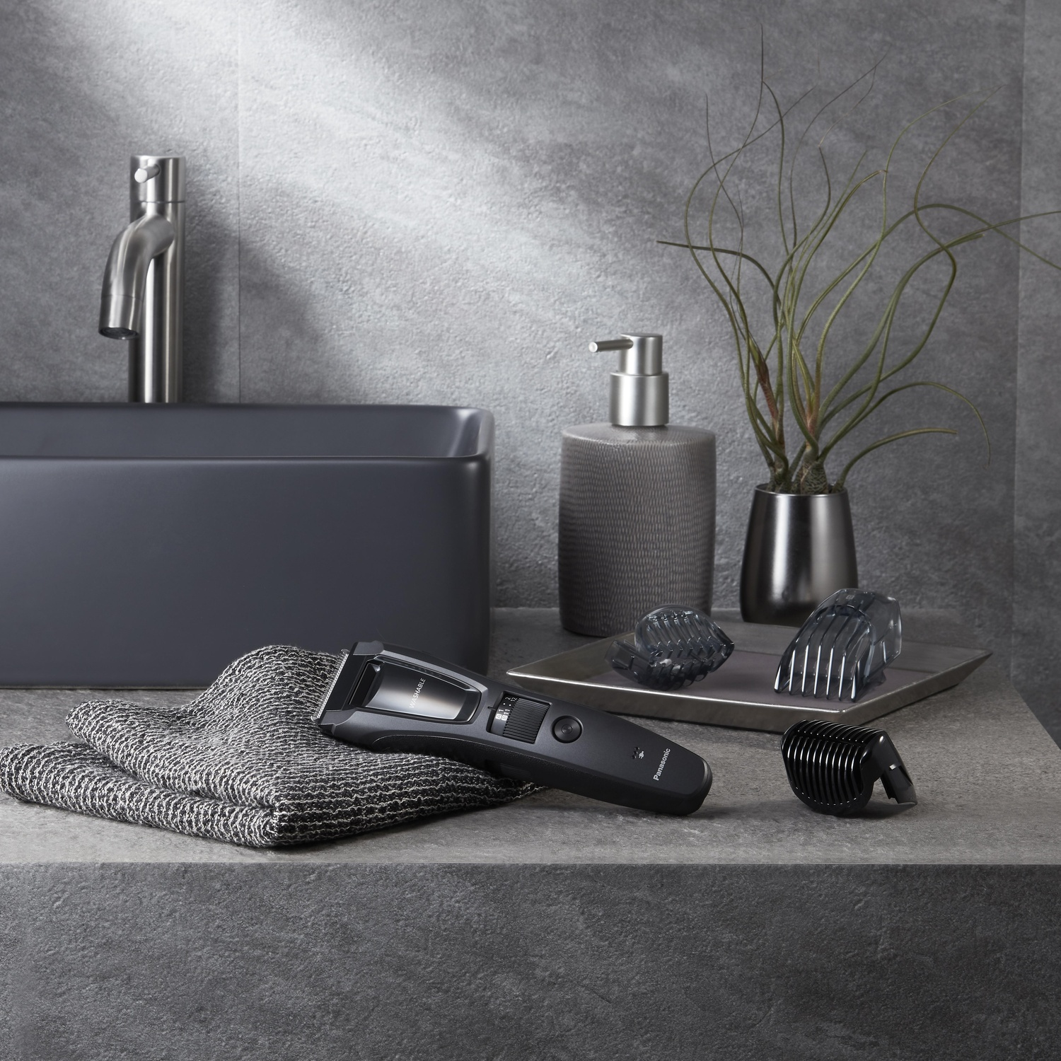 Allestimento bagno: lavandino nero, kit rasatura, dispenser sapone, pianta e accessori su superficie grigia.