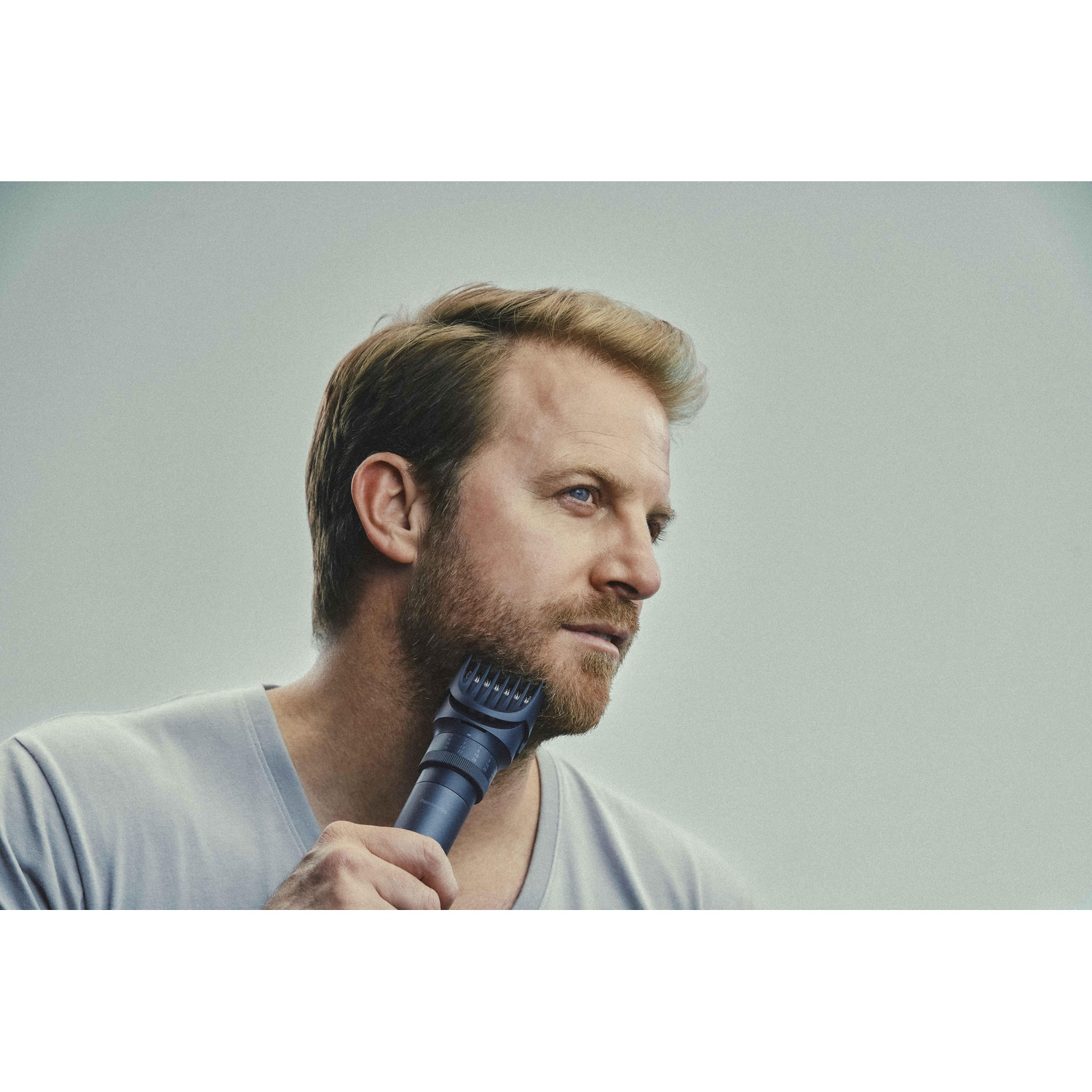 Uomo con la barba che si rade la barba con un regolabarba blu, che guarda altrove.