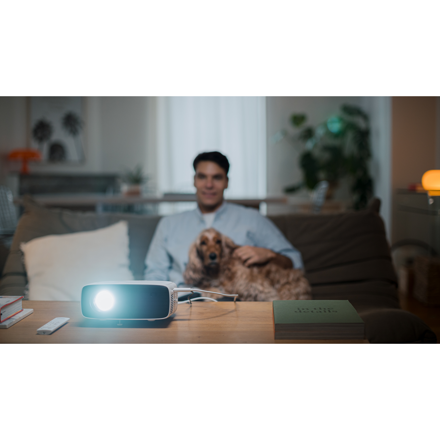 Un homme avec un chien sur un canapé, projecteur sur une table. Le projecteur brille. Livres et télécommande sur la table.