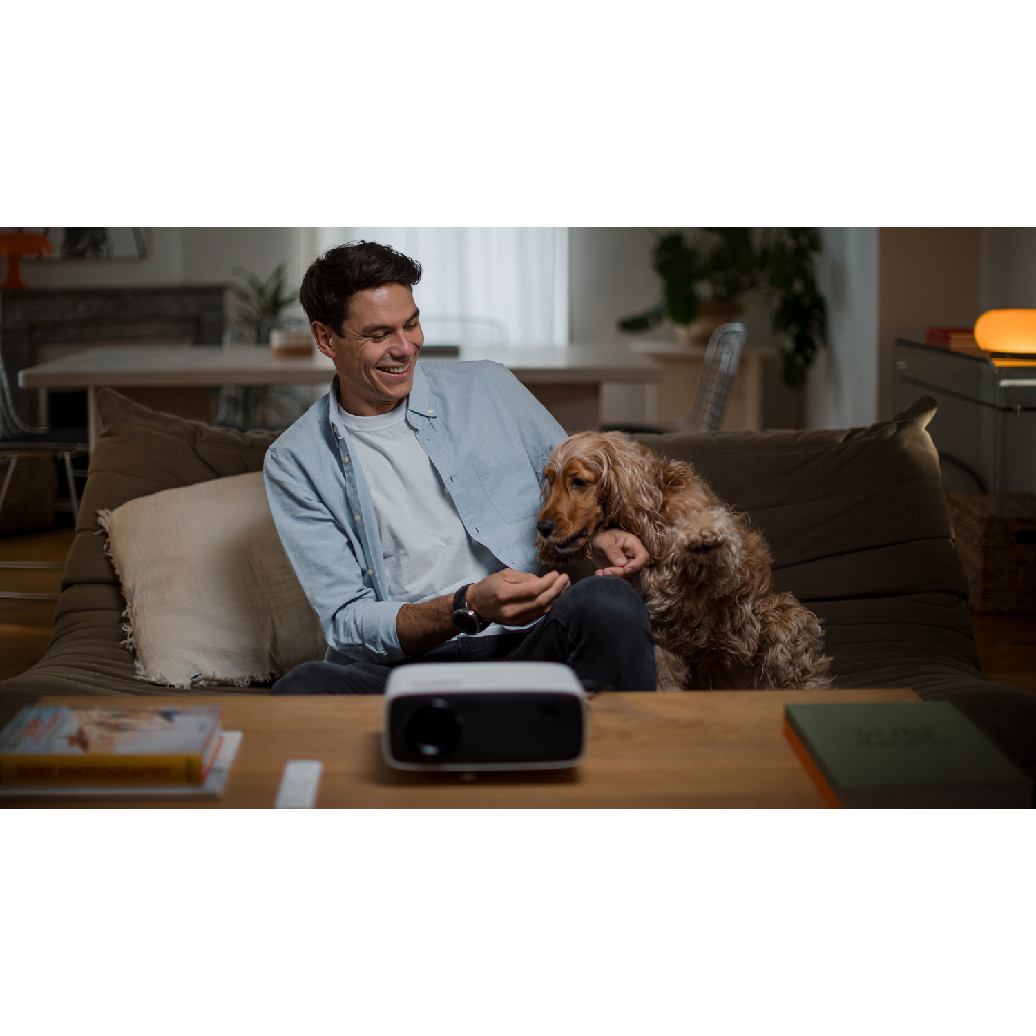 Un homme sourit, caressant un chien brun sur un canapé. Un projecteur est sur la table. Livres et décor au fond.