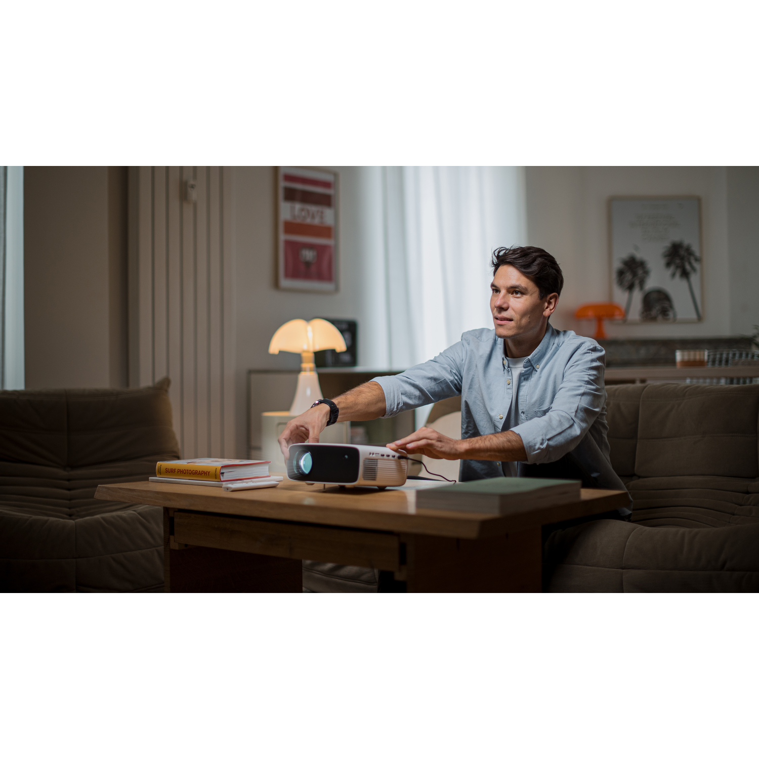 Un homme ajuste un projecteur sur une table. Livres et canapé au premier plan. Art et lampe en arrière-plan.