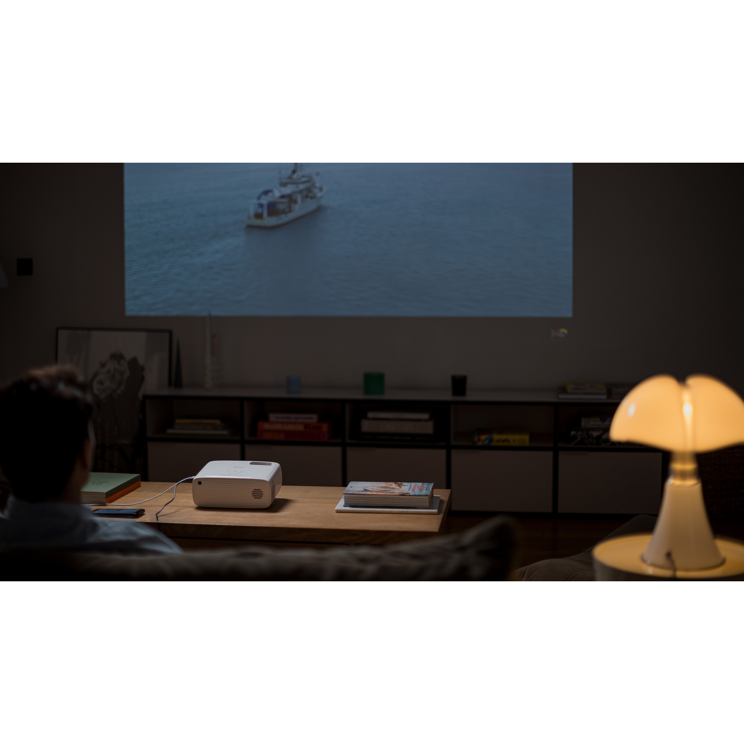 Un homme regarde un film projeté sur le mur. Un projecteur est sur la table. Une lampe blanche est à droite.