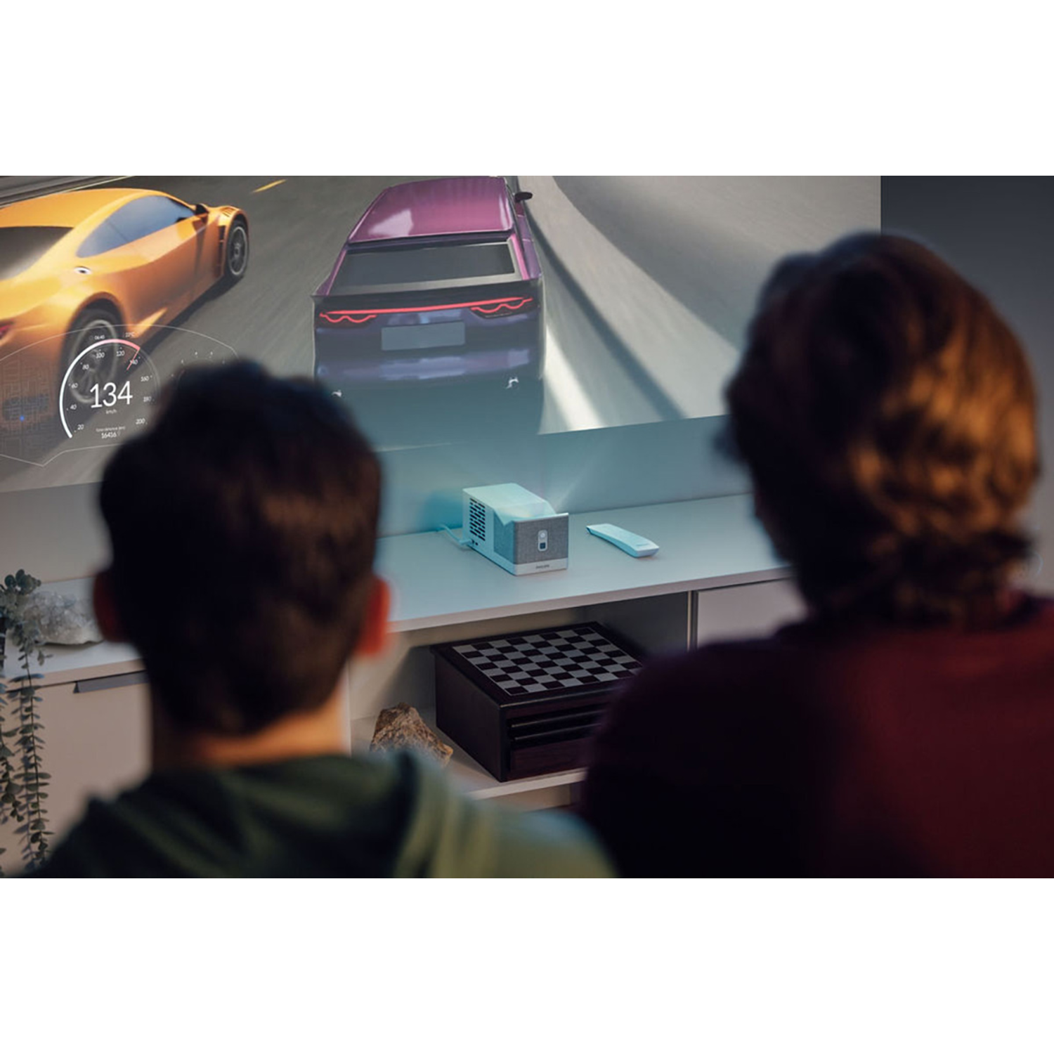 Deux personnes regardent un jeu de course de voitures projeté. Un projecteur, une télécommande et un jeu d'échecs sont sur une étagère.