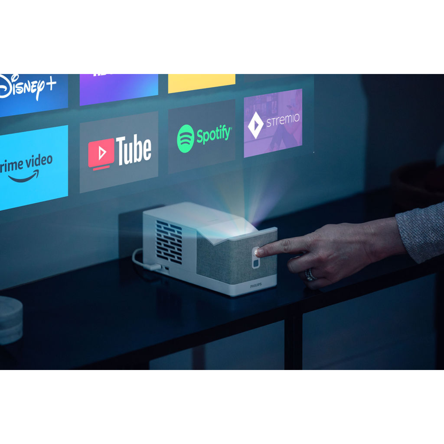 Une main appuie sur un bouton d'un projecteur Philips projetant des logos d'applications sur un mur : Disney+, YouTube, Spotify et Stremio.