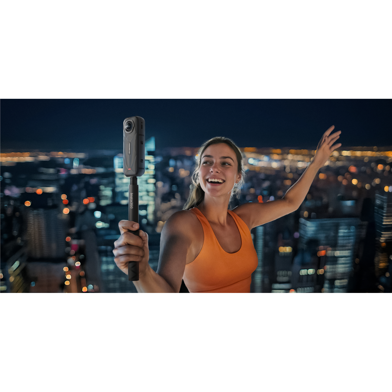 Une femme avec un haut orange tenant un bâton de caméra 360 souriant avec un paysage urbain en arrière-plan.
