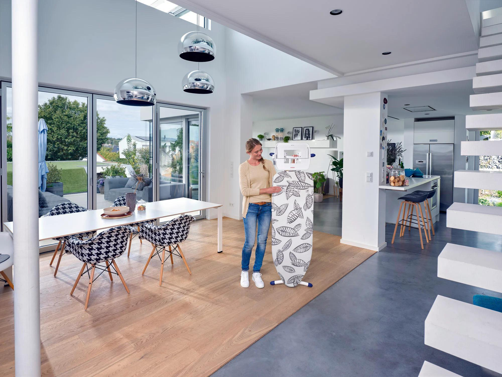 Frau mit Bügelbrett in modernem Wohnraum mit großen Fenstern und weißer Treppe.