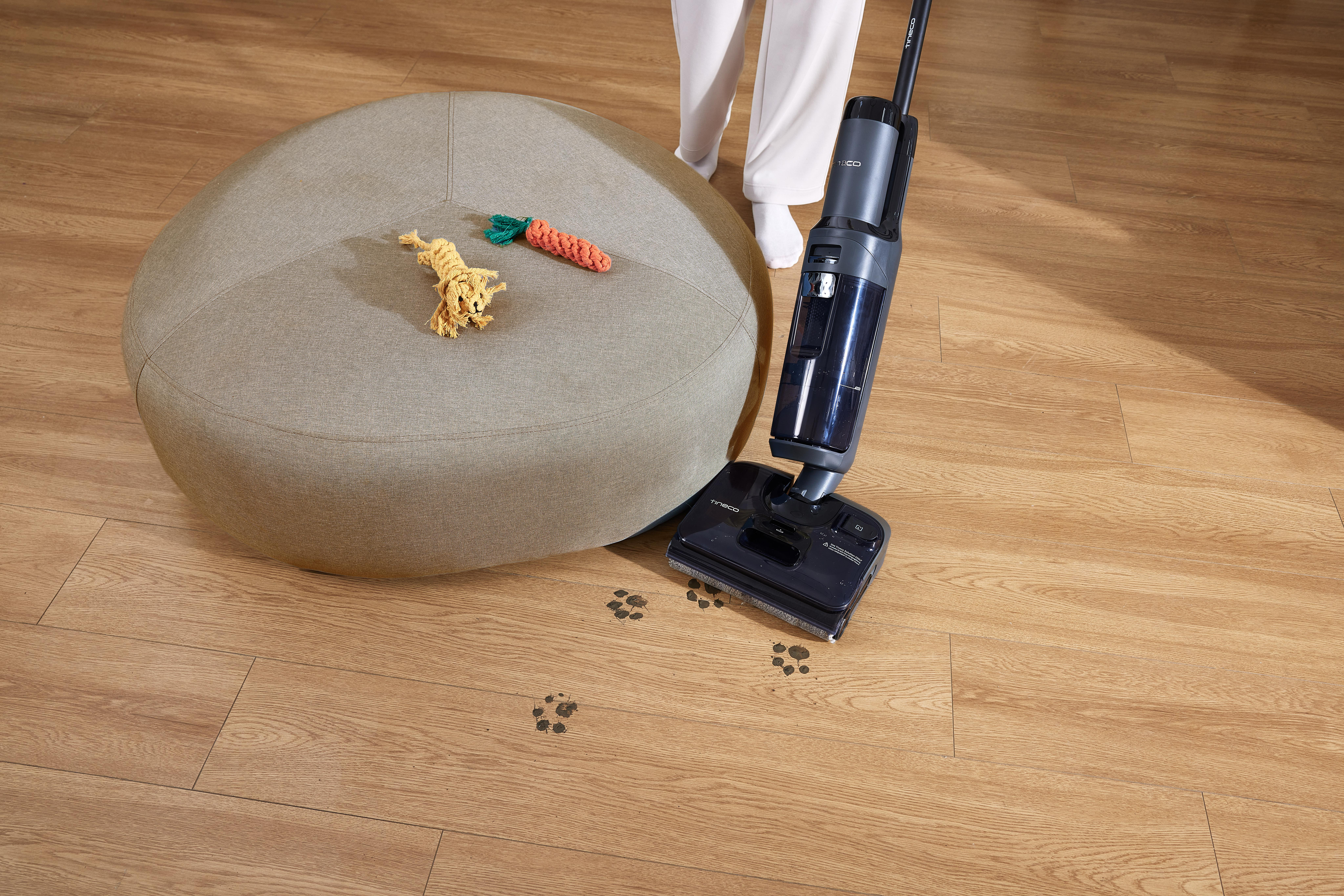 Person saugt Holzboden mit einem kabellosen Staubsauger, in der Nähe von Kissen und Spielzeug.