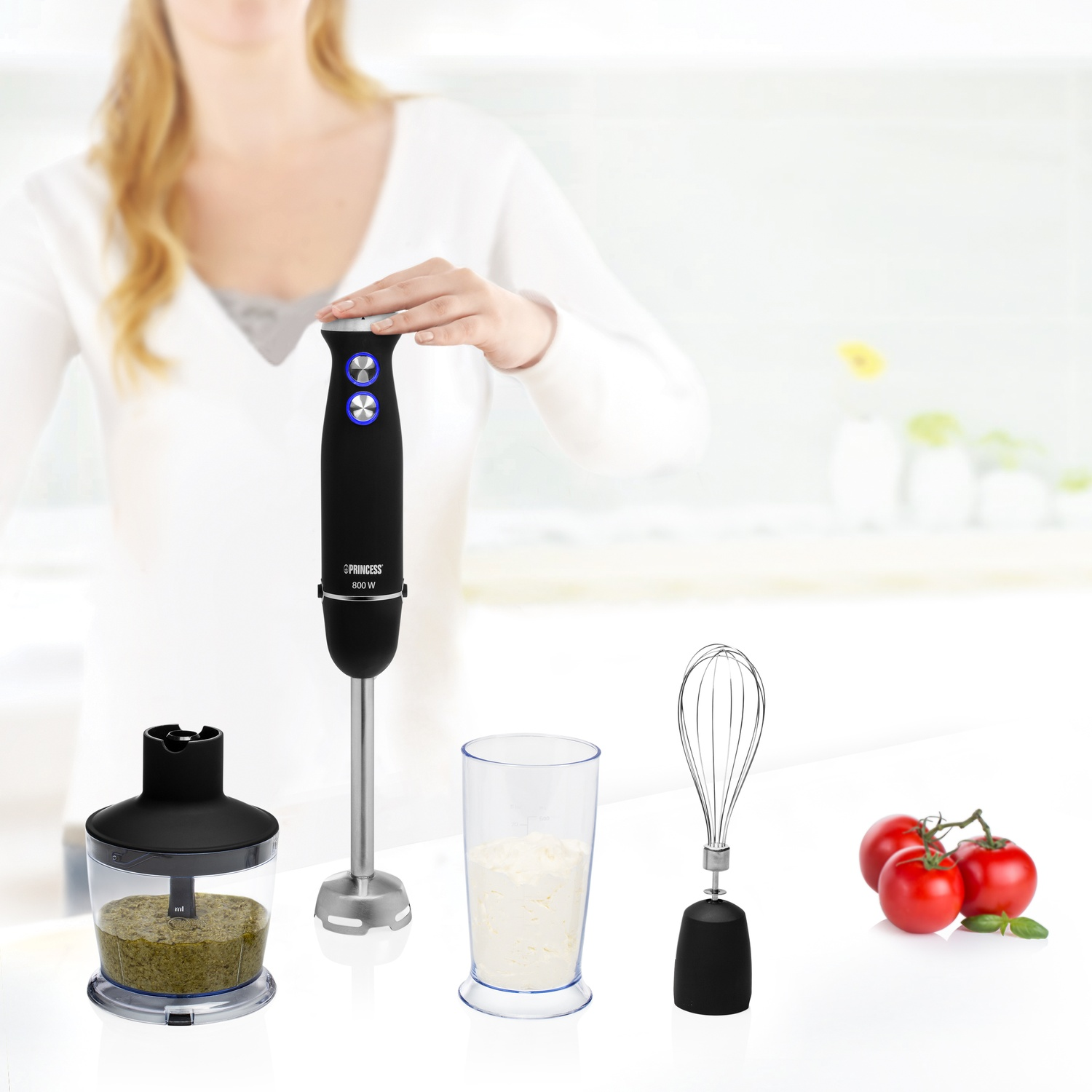 Un frullatore a immersione nero con accessori, tra cui un contenitore con contenuto verde e pomodori.