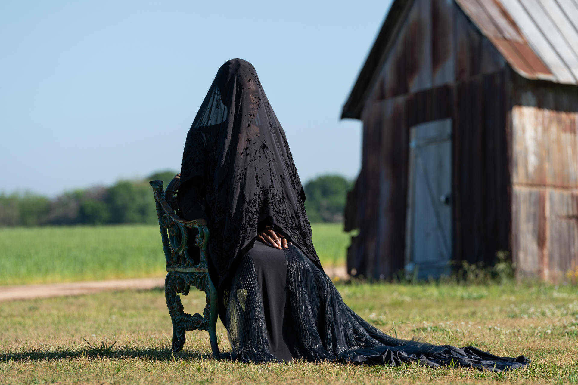 Person mit schwarzem Schleier sitzt auf einer Bank auf einem Feld, Scheune im Hintergrund.