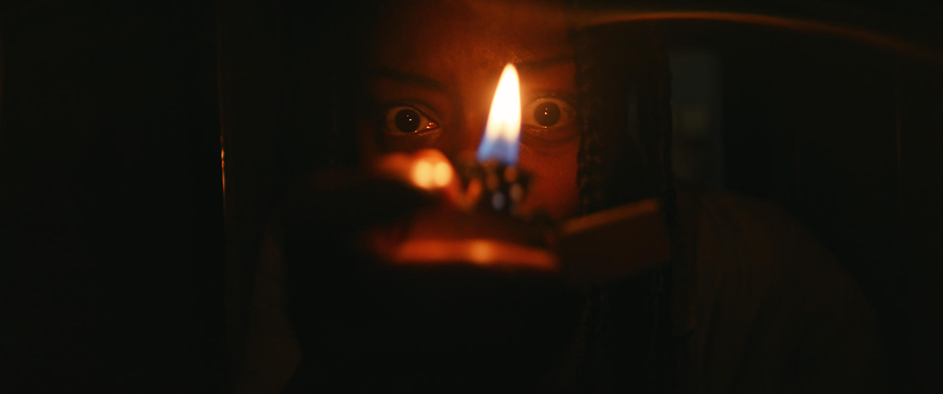 Nahaufnahme eines Gesichts, beleuchtet von einer Feuerzeugflamme, mit weit aufgerissenen Augen in dunkler Umgebung.