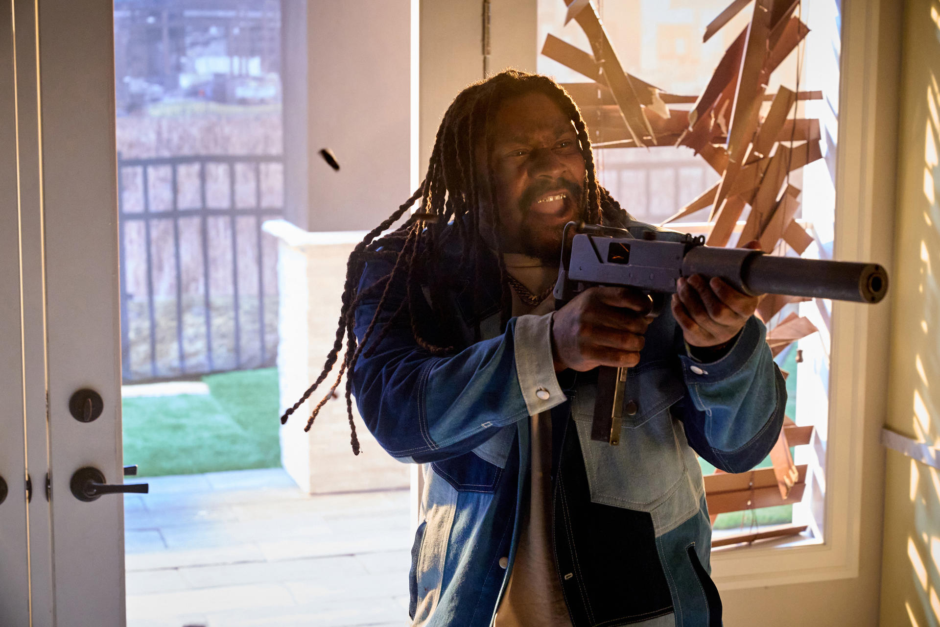 Ein Mann mit Dreadlocks hält eine Waffe und schaut wütend. Die Szene scheint sich in einem Innenraum in der Nähe eines Fensters abzuspielen.