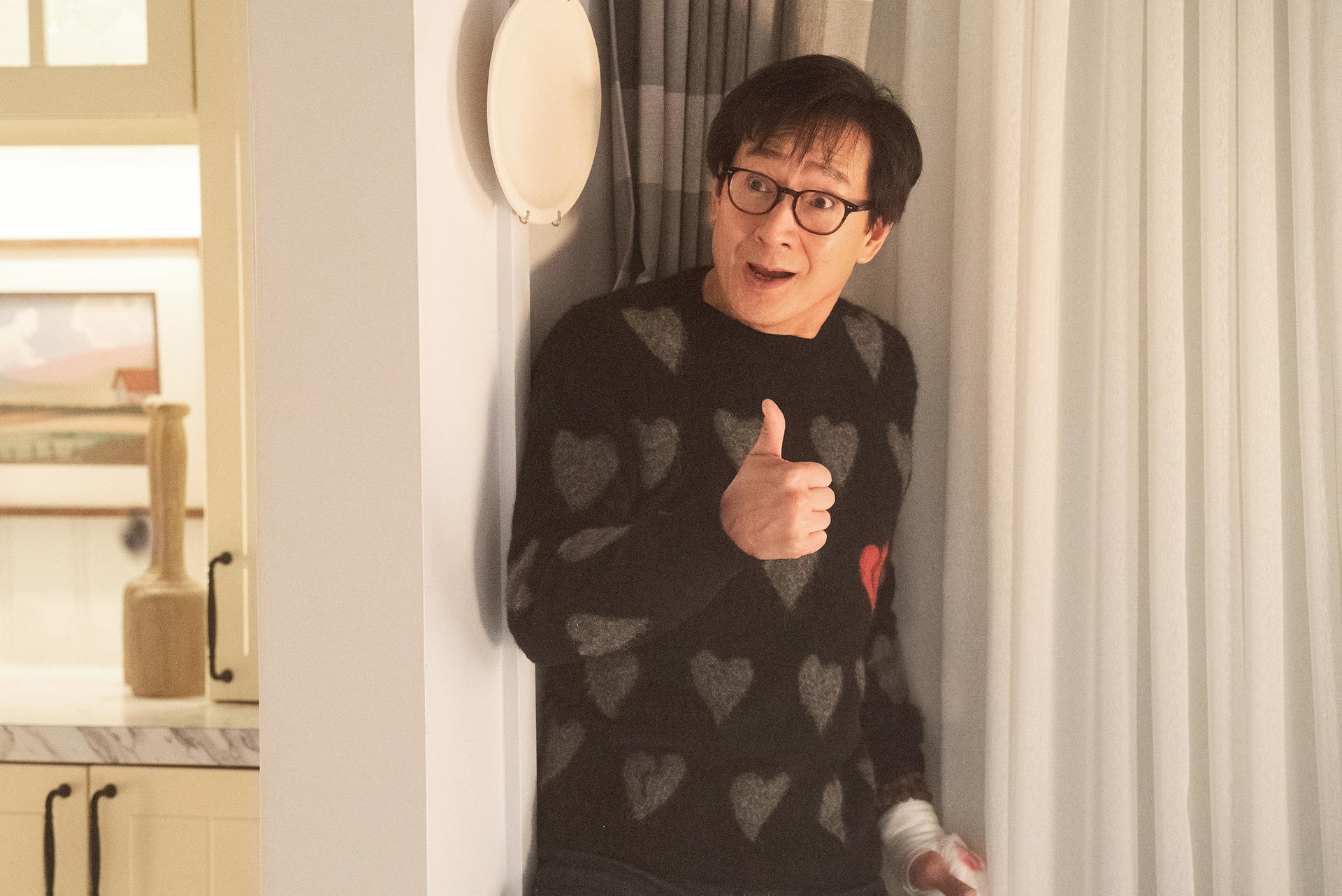 Mann mit Brille gibt Daumen hoch, lehnt sich in einen Türrahmen. Er trägt einen Pullover mit Herzmuster.