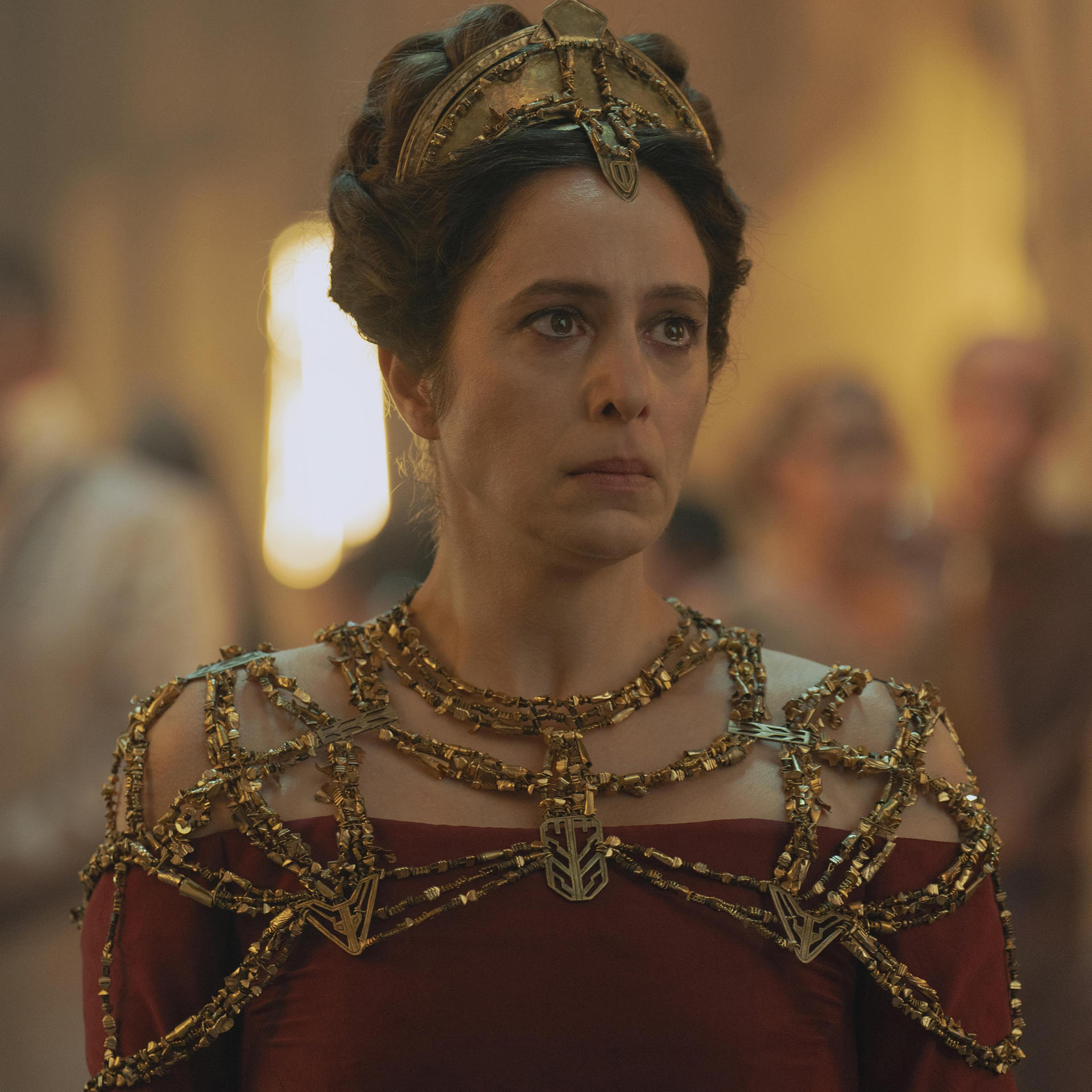 Frau mit goldenem Kopfschmuck und Halskette, rotes Kleid, ernst blickend, verschwommener Hintergrund.