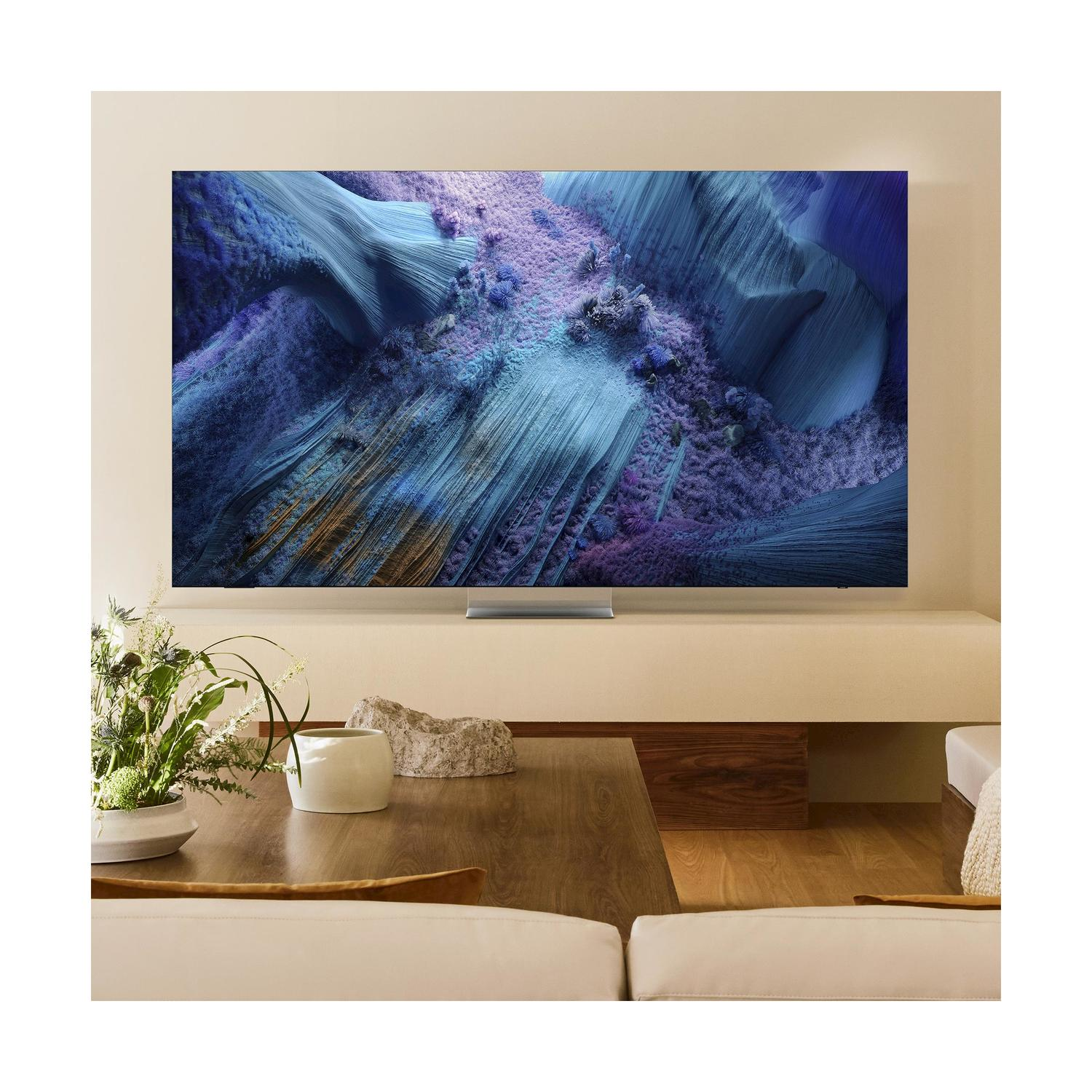 TV montata a parete, che mostra un paesaggio astratto. Tavolo in legno, vaso e decorazioni.