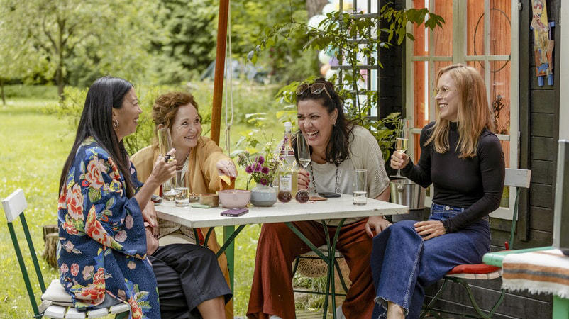 Vier Frauen lachen an einem Tisch im Freien. Es gibt einen Tisch mit Gläsern und etwas Essen.