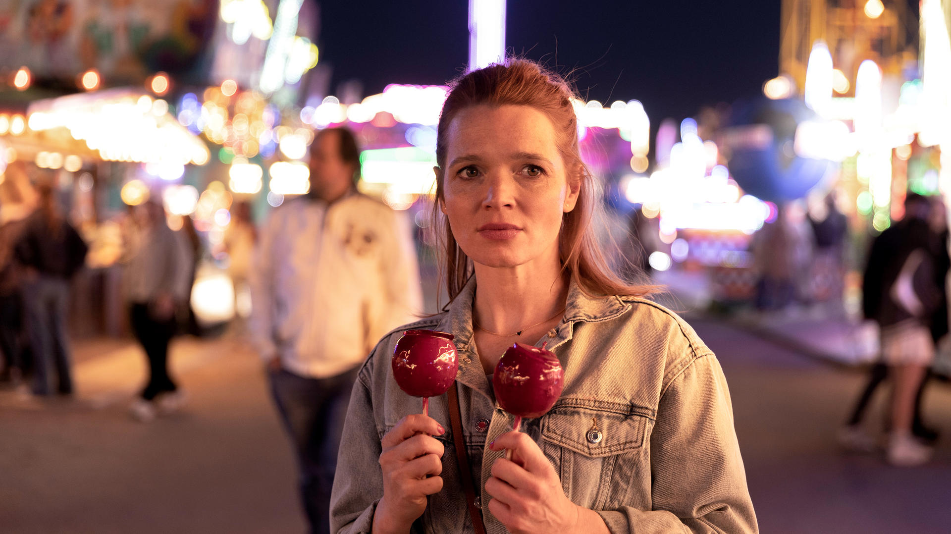 Frau auf einem Jahrmarkt mit zwei Zuckeräpfeln; unscharfe Lichter im Hintergrund.