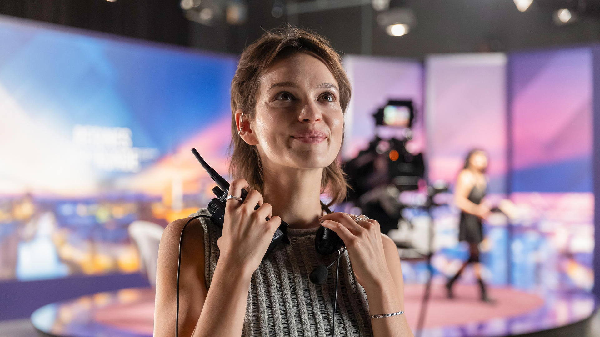 Frau im Studio, Headset und Mikrofon haltend, lächelnd. Hintergrund mit Bildschirmen.