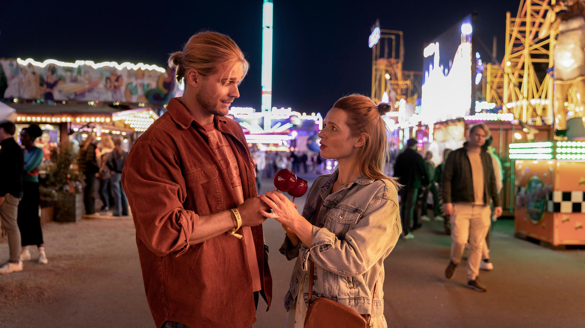 Paar auf dem Jahrmarkt, Mann gibt Frau einen kandierten Apfel. Hintergrund: Lichter, Stände, Nacht.