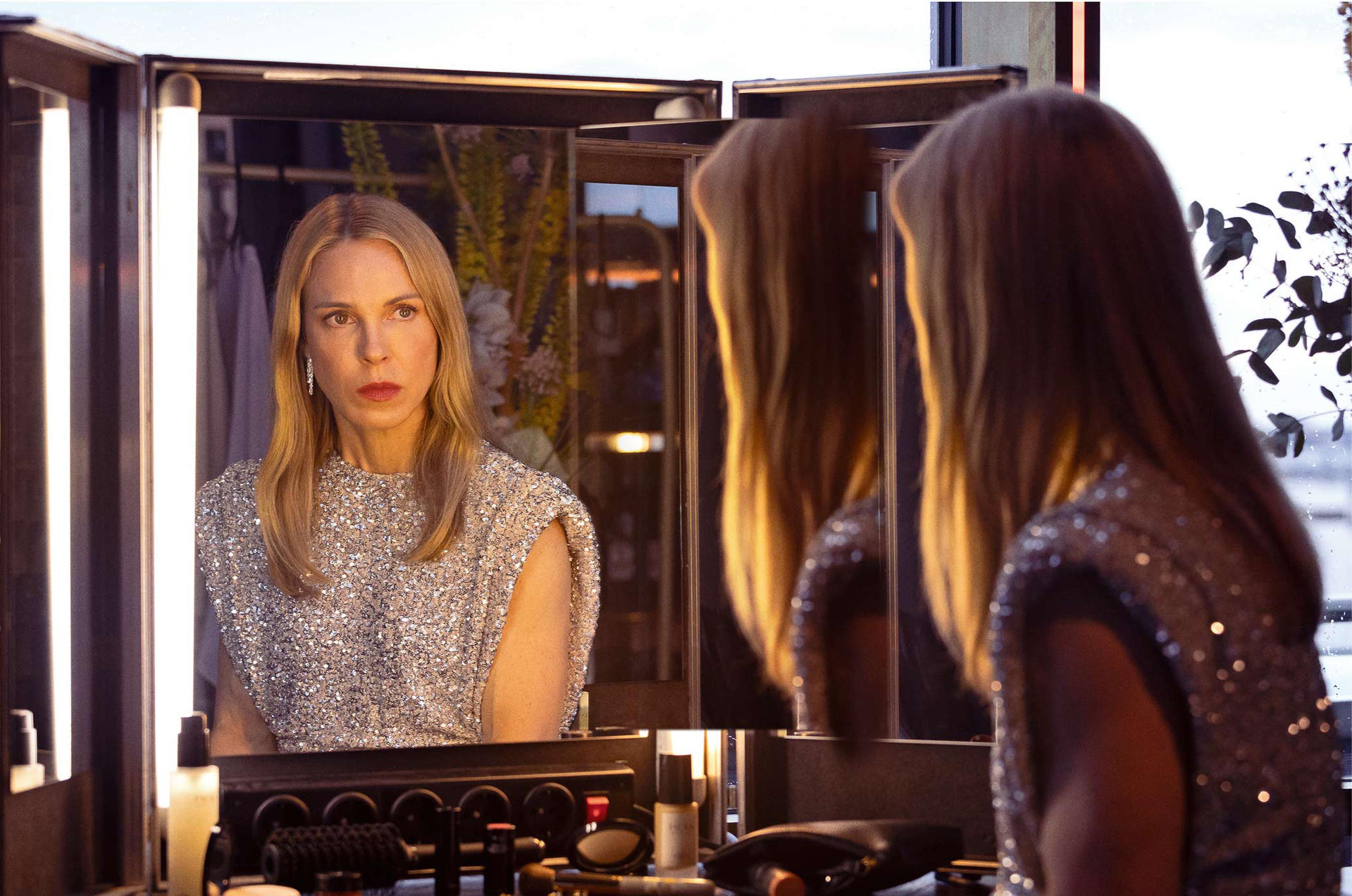 Frau im Pailletten-Top blickt in einen Spiegel. Spiegelungen zeigen ihr Haar und einen Blick aus dem Fenster. Make-up und Lichter rahmen den Spiegel ein.