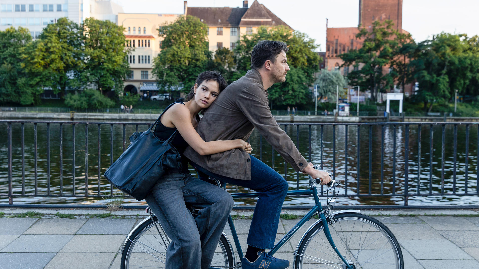 Ein Paar auf einem Fahrrad am Fluss, die Frau umarmt den Mann. Grüne Bäume und Gebäude.