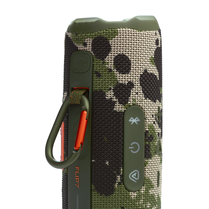 Un altavoz JBL Flip 7 con un patrón de camuflaje verde y marrón y detalles en naranja.