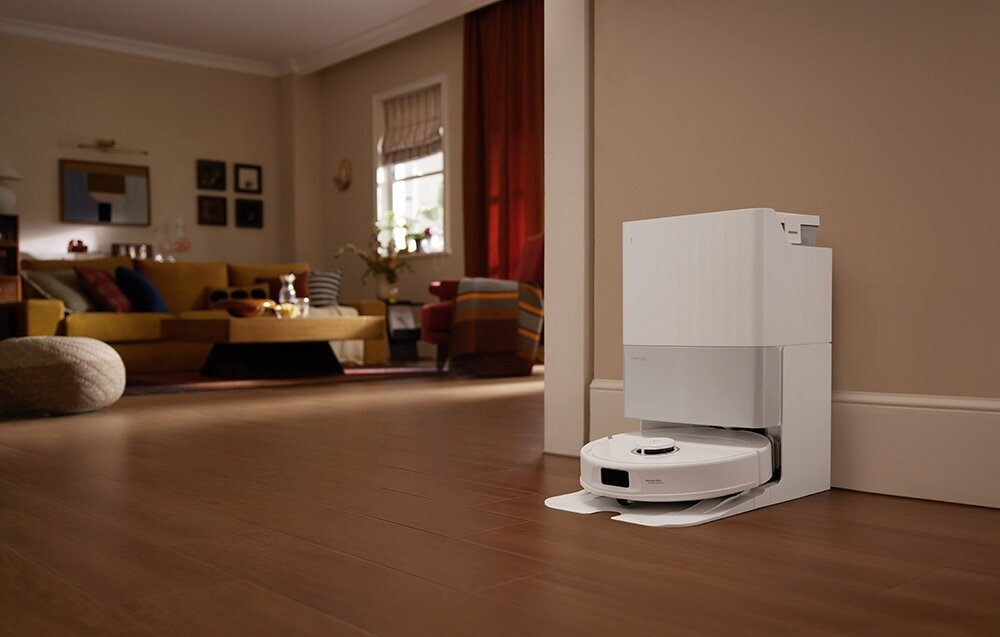Robot odkurzacz jest zadokowany, z białą stacją ładowania i czyszczenia. Tło salonu.
