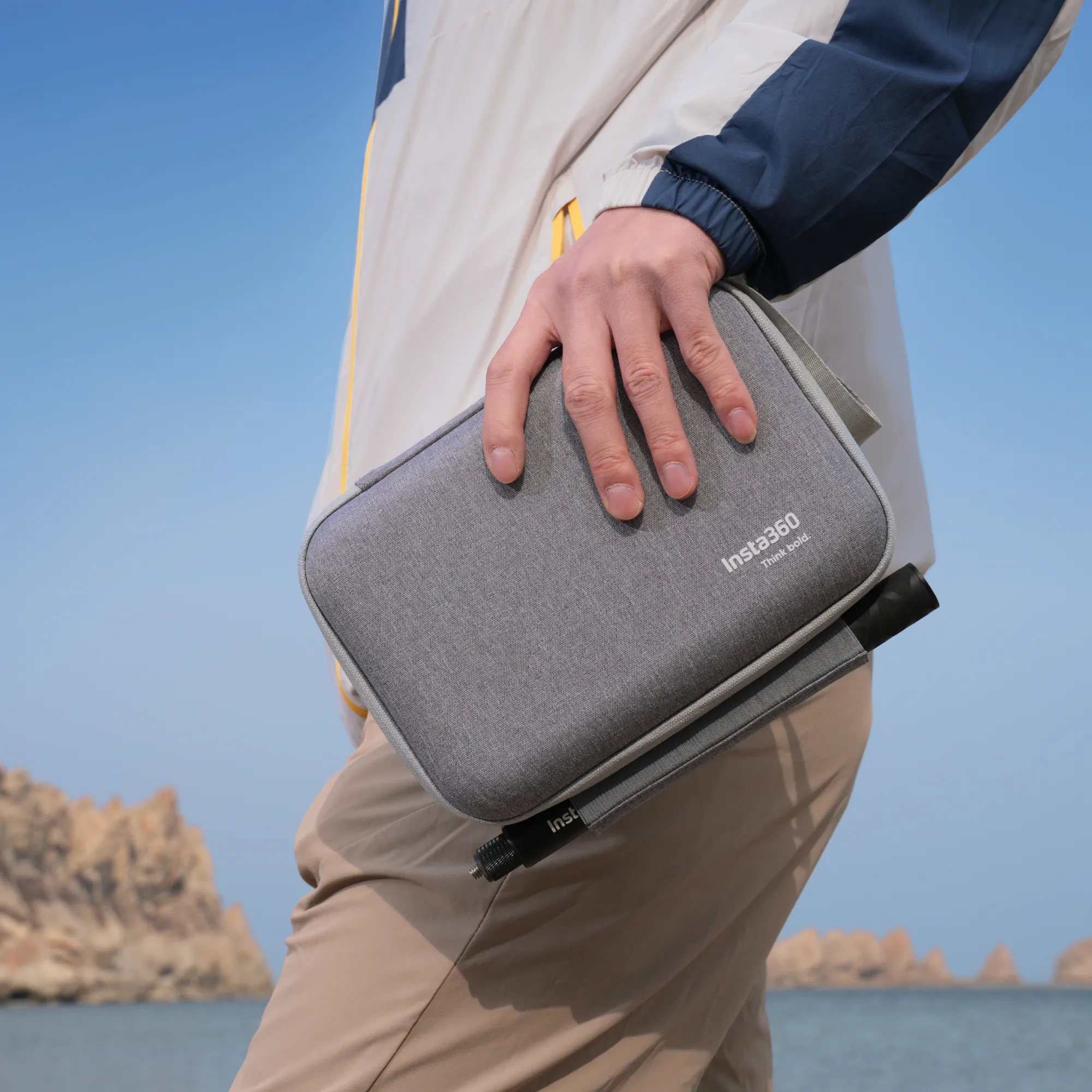 Person hält eine graue Tasche mit Kamera und Zubehör. Blauer Himmel und Felsen im Hintergrund.