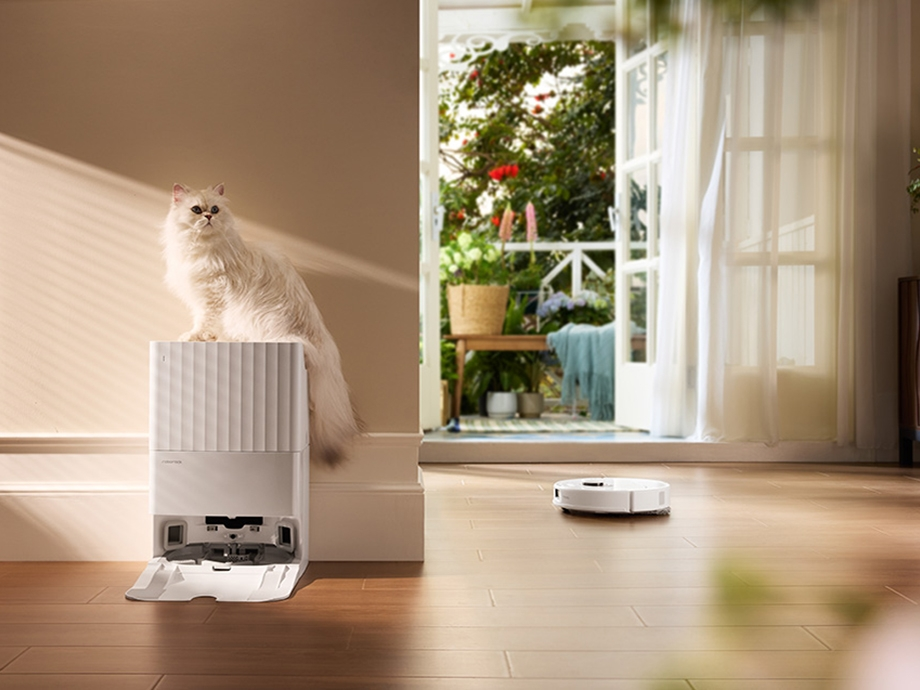 Biały kot siedzi na białym odkurzaczu, obserwując robota sprzątającego.