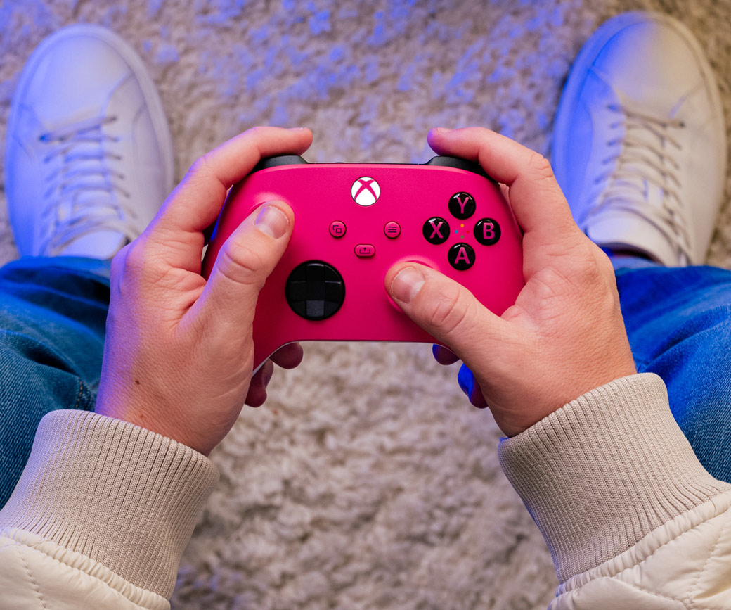Hände halten einen rosa Xbox-Controller. Person trägt Jeans und weiße Schuhe auf einem Teppich.
