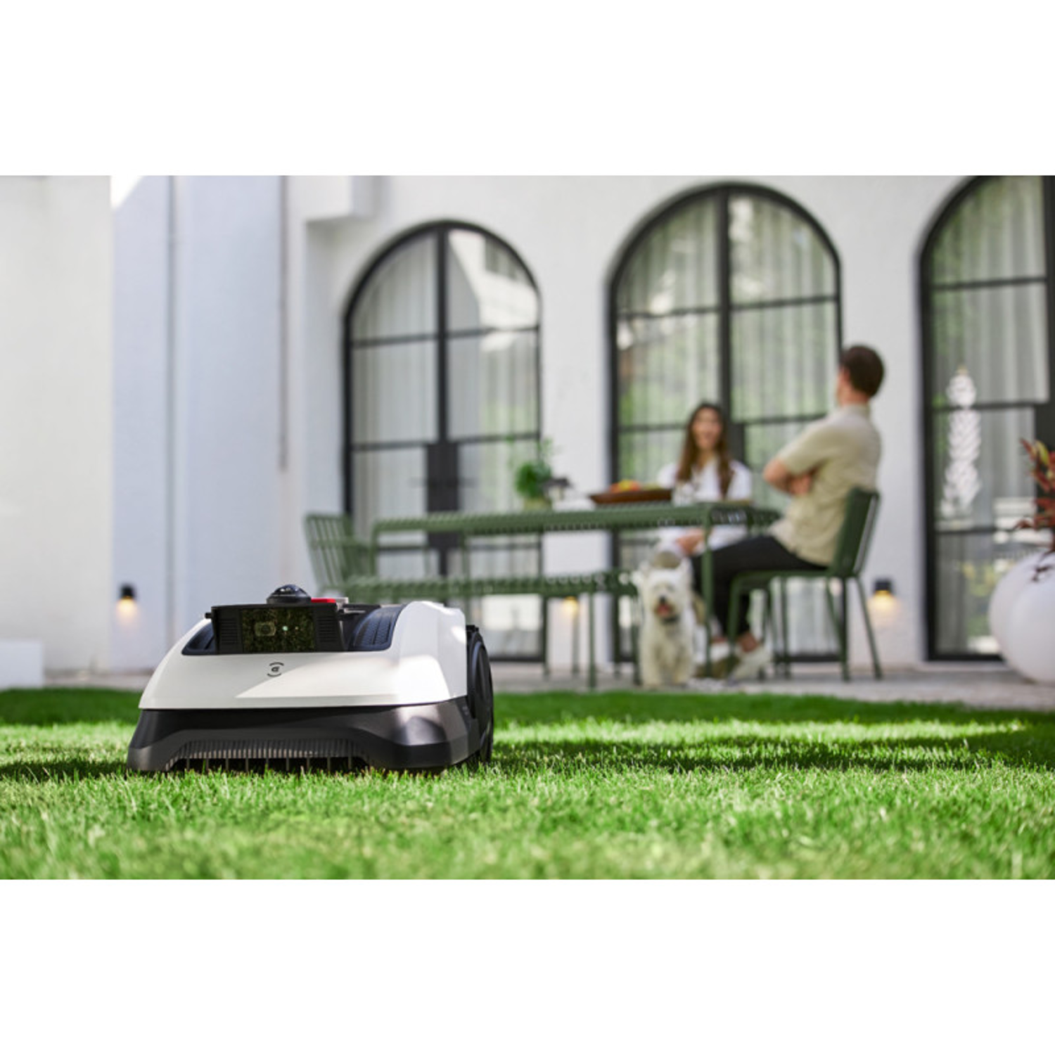Robot lawnmower on green grass with people and dog in background.