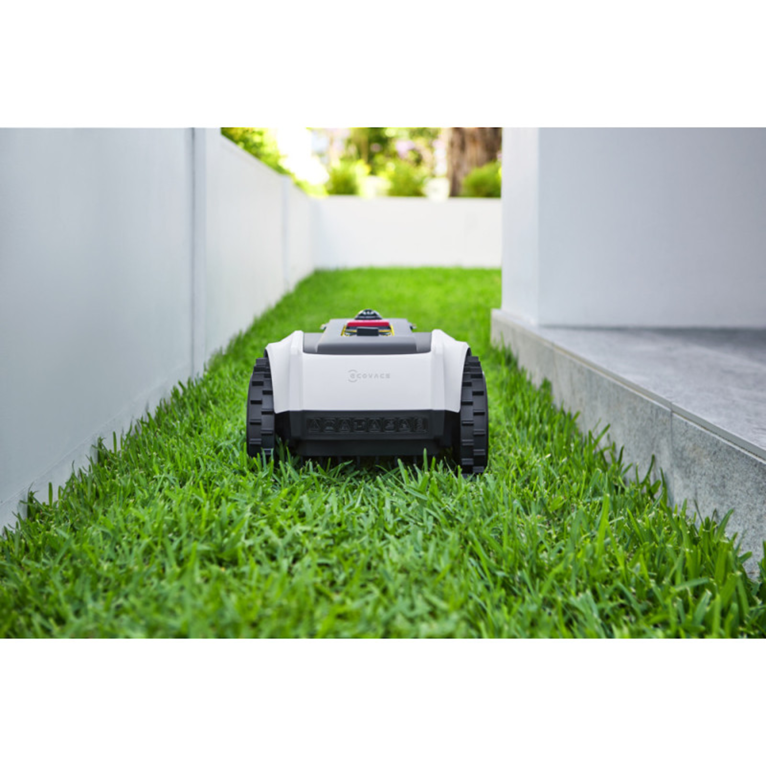 White robotic lawnmower on green grass, moving through a narrow passage.
