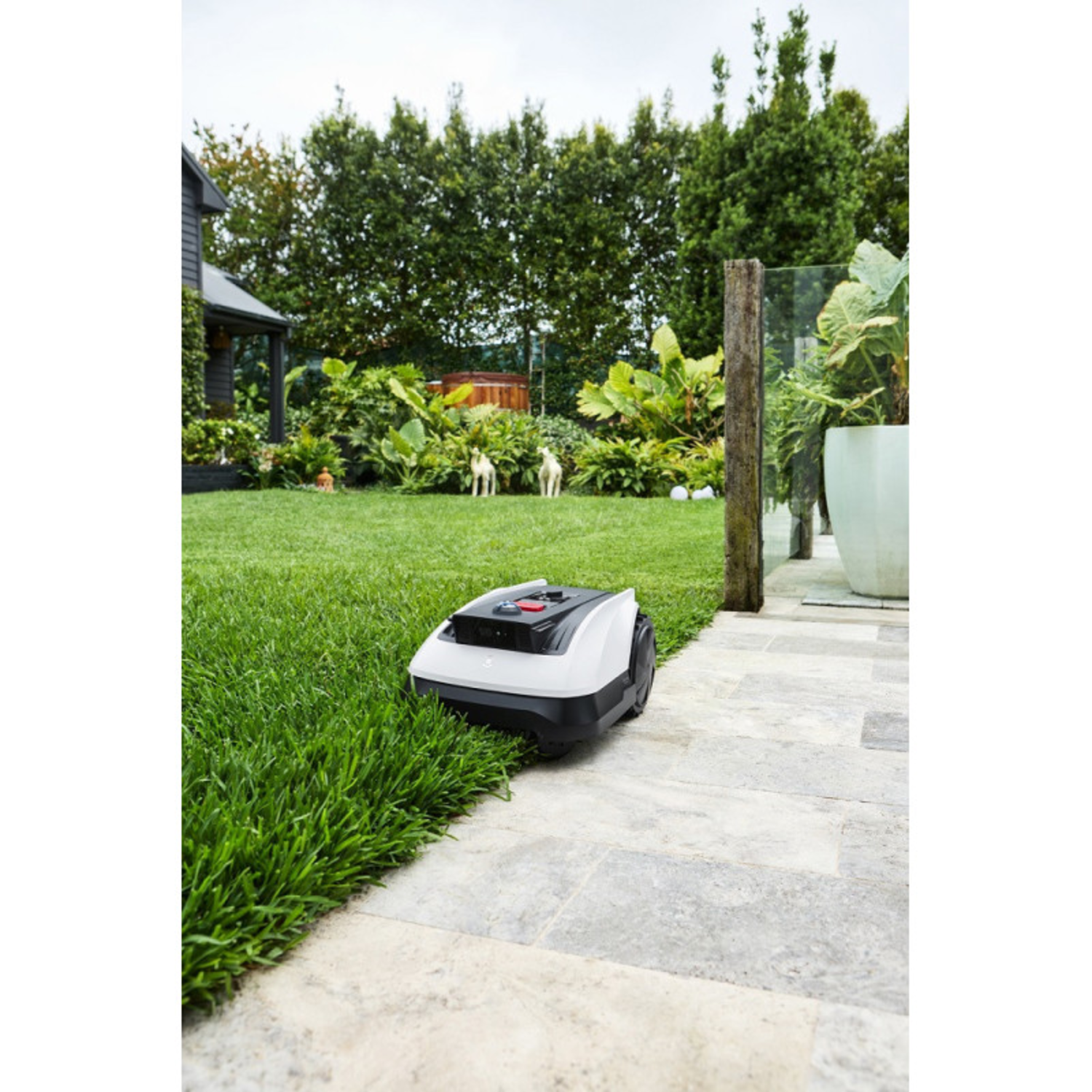 Robot lawn mower on a grassy lawn next to a stone walkway. Trees in the background.