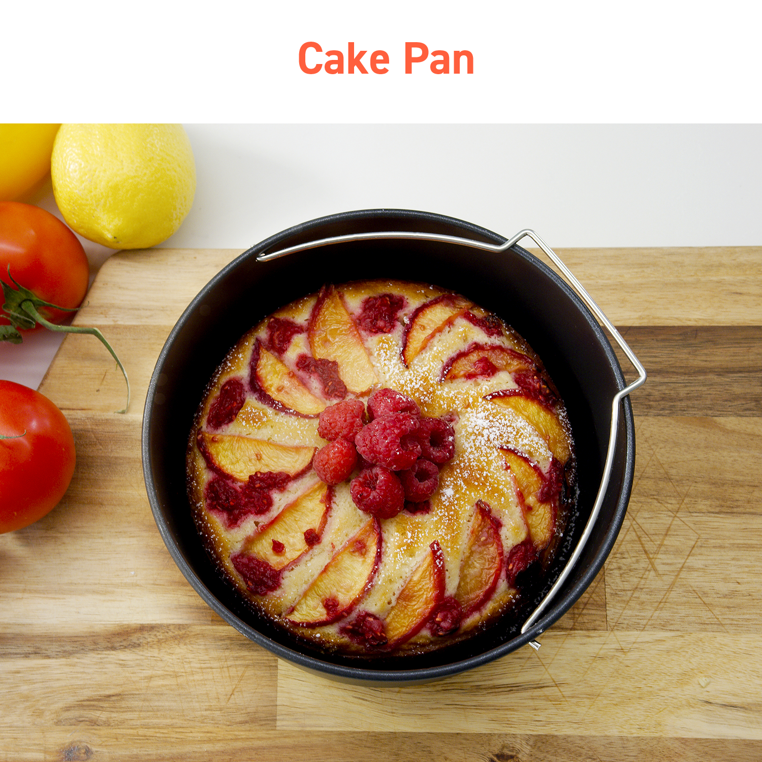 Cake pan with a cake, raspberries, and peaches on a wooden surface. The pan has a handle.
