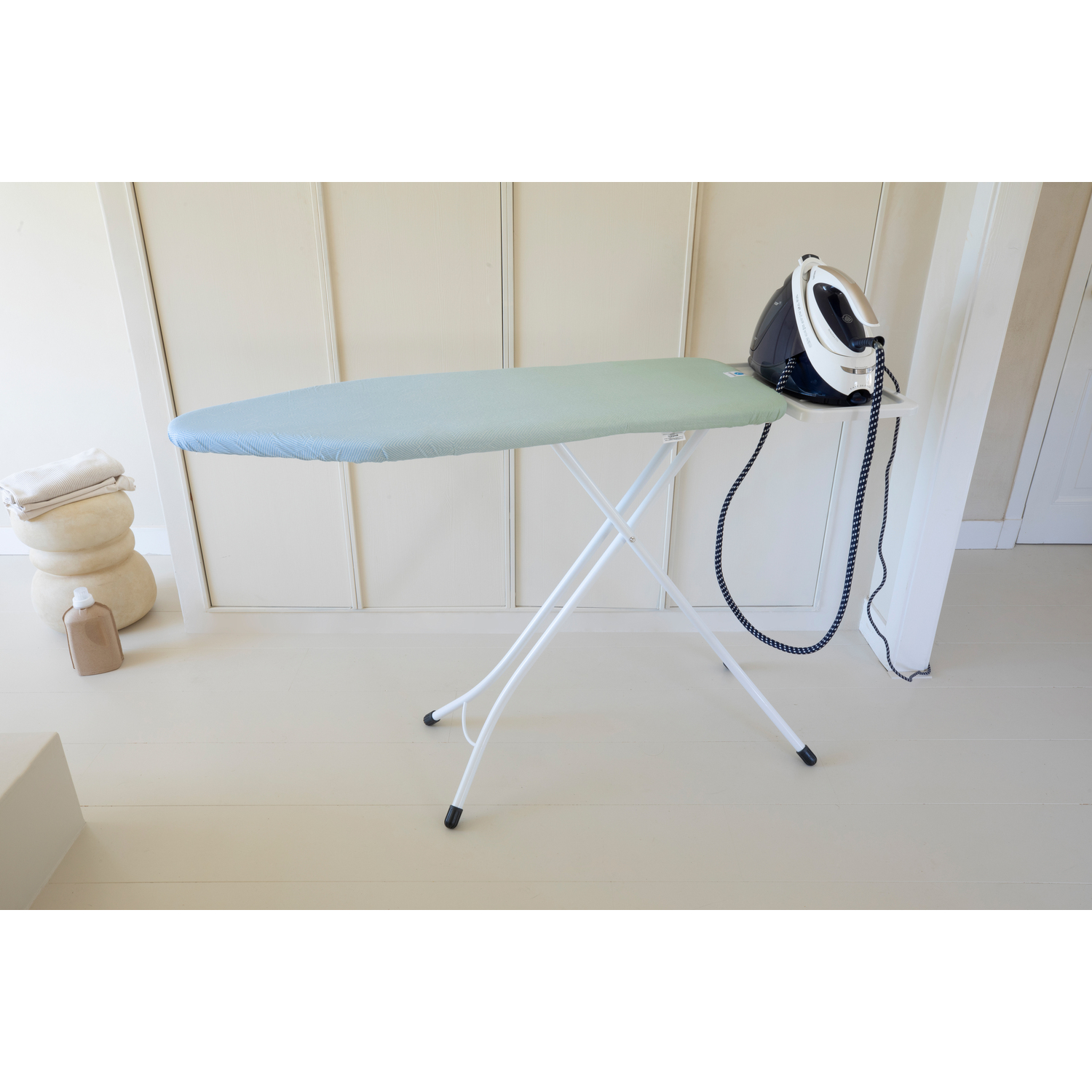 Ironing board with a light blue cover, iron, and other items on a white floor.