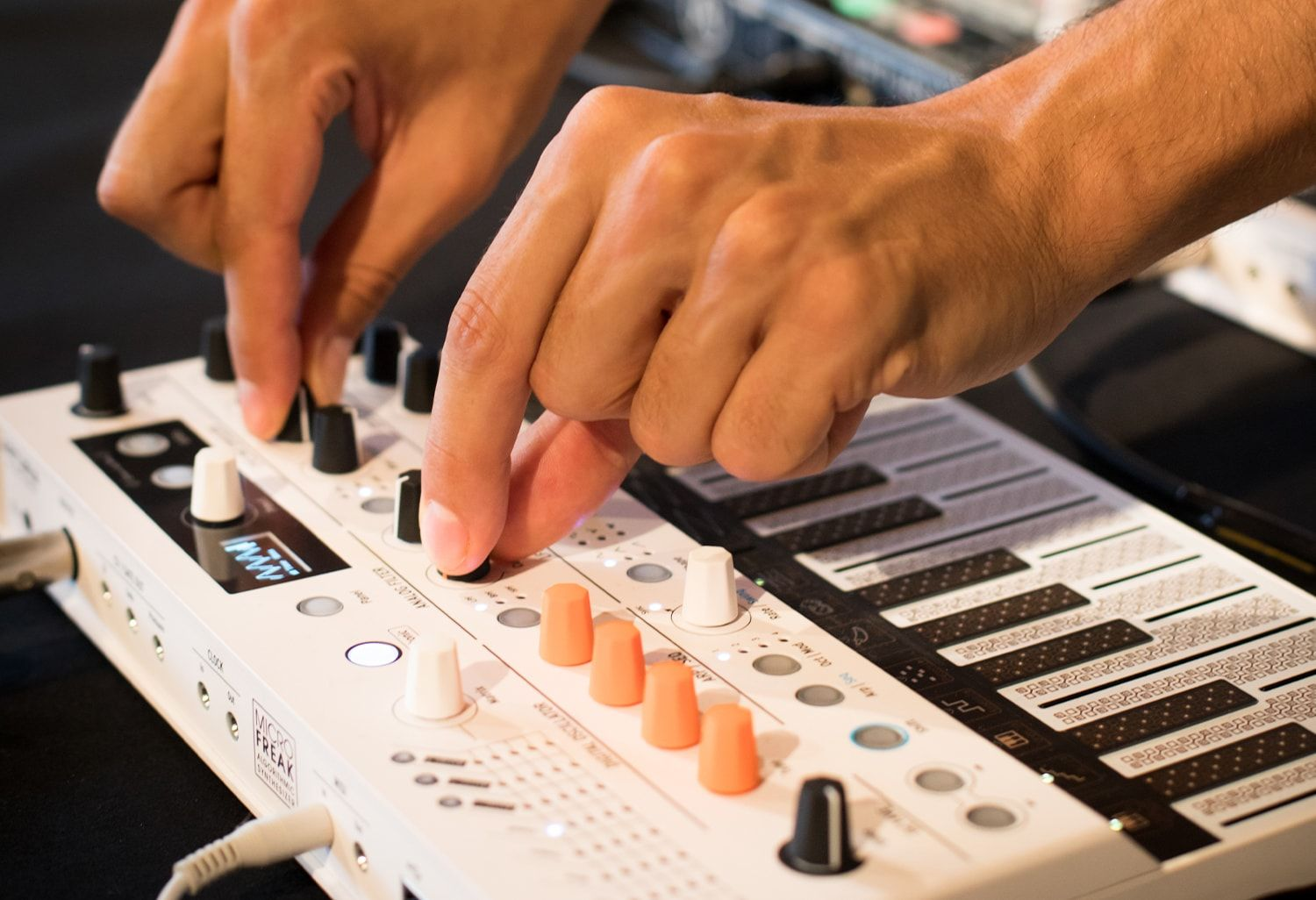 Mains réglant des boutons sur un synthétiseur blanc. Divers boutons et touches sont visibles.