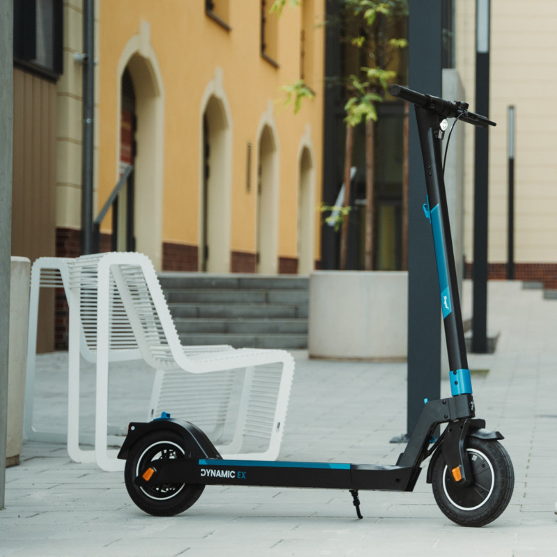 Hulajnoga elektryczna zaparkowana na chodniku. Biała ławka i budynek w tle.