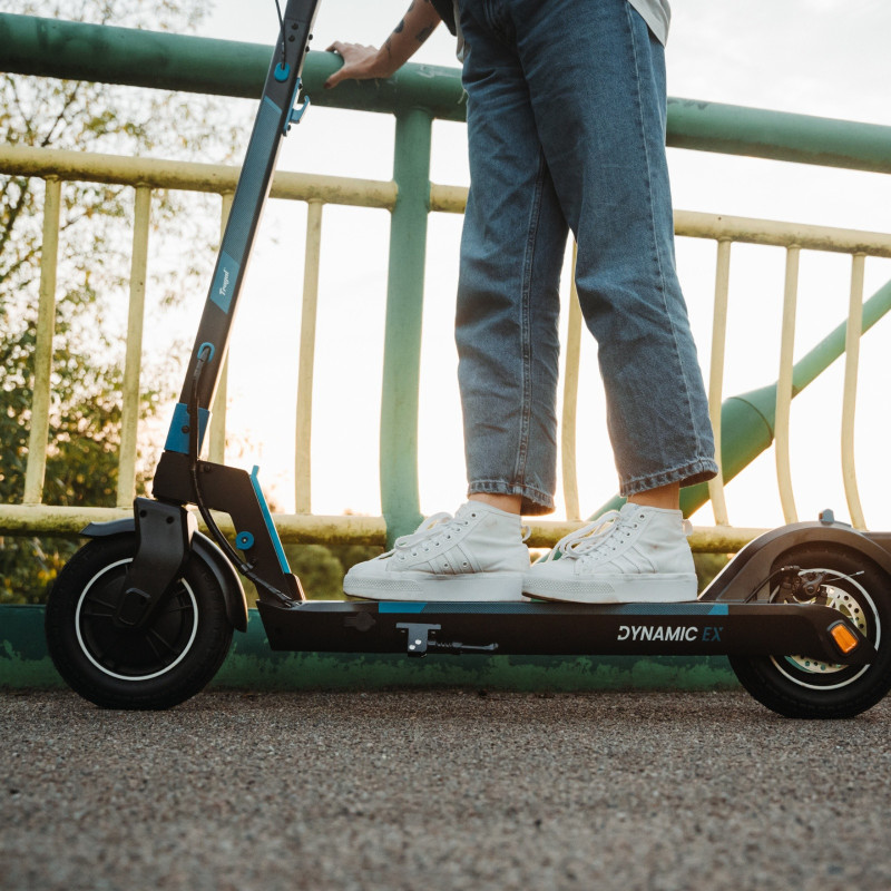 Osoba jadąca czarno-niebieską hulajnogą na moście. Słoneczny dzień.