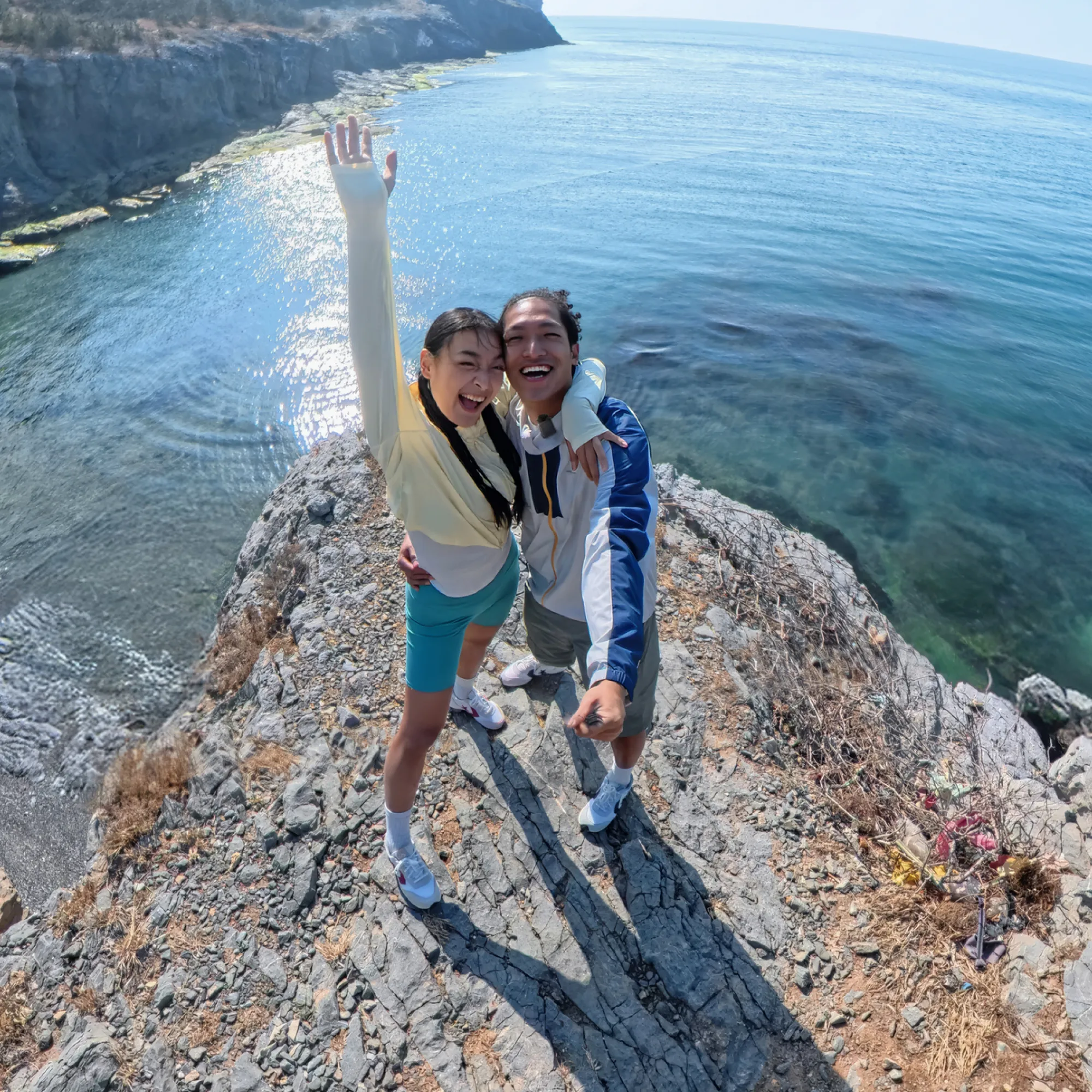 Zwei lächelnde Personen, eine mit erhobenem Arm auf einer Klippe mit Blick auf das Meer, sonniger Tag.