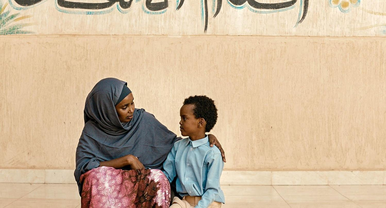 Eine Frau mit Hijab tröstet einen Jungen. Sie sitzen zusammen, eine Wand mit Schrift im Hintergrund.